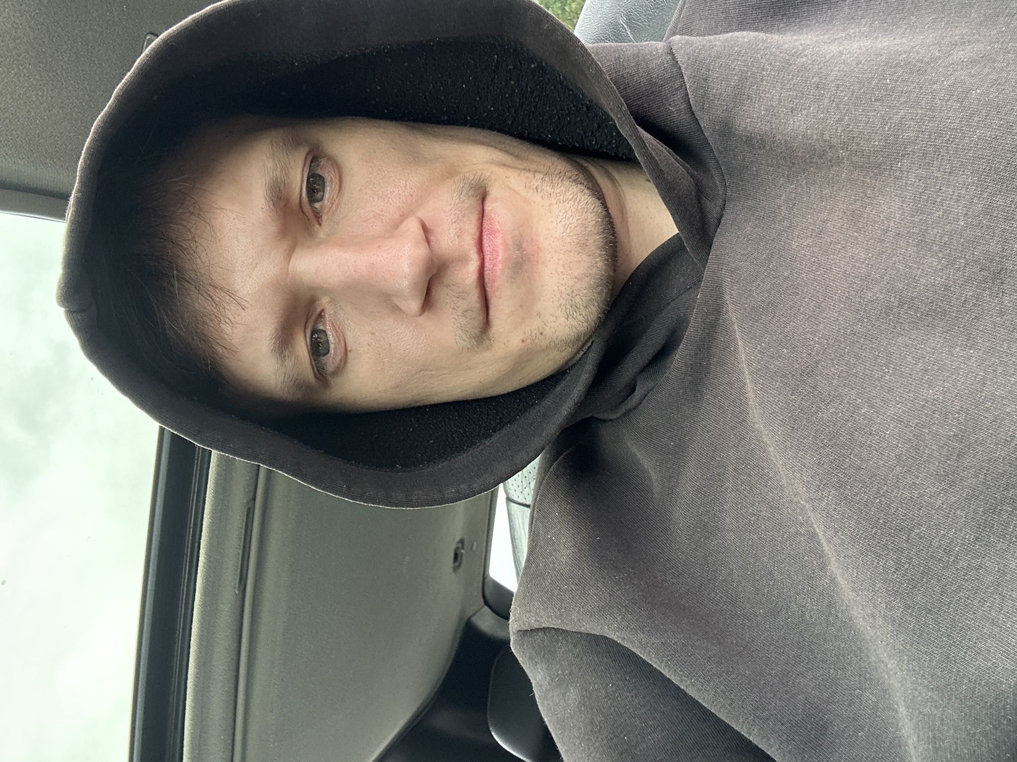 A man wearing a dark hoodie is seen from the chest up, inside a vehicle. The man has light skin, short brown hair, and is looking directly at the camera. The interior of the car, including the headliner and a sunroof, is visible. The background shows a cloudy sky through the sunroof. The image appears to be a selfie taken by the man. The specific location of Makiv, Ukraine, is not determinable from the image content.