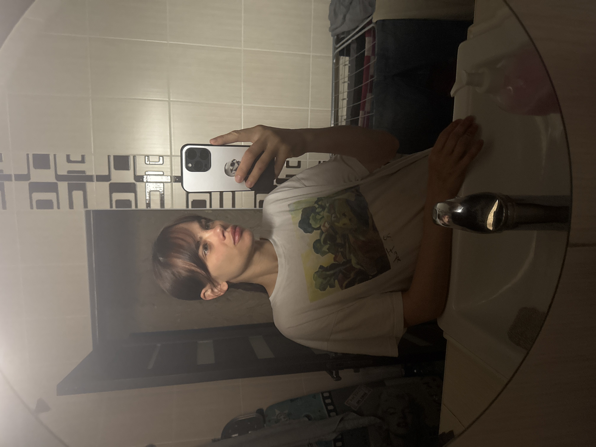 A person is taking a selfie in a bathroom mirror. The person has light skin, brown hair with bangs, and is wearing a white t-shirt with a graphic design. The person is holding a silver smartphone with a triple-lens camera. The bathroom has white and grey tiled walls, a white sink, and a chrome faucet. A metal rack with clothes is visible in the background. There is no evidence to suggest this photograph was taken in Rostov-on-Don, Russia.