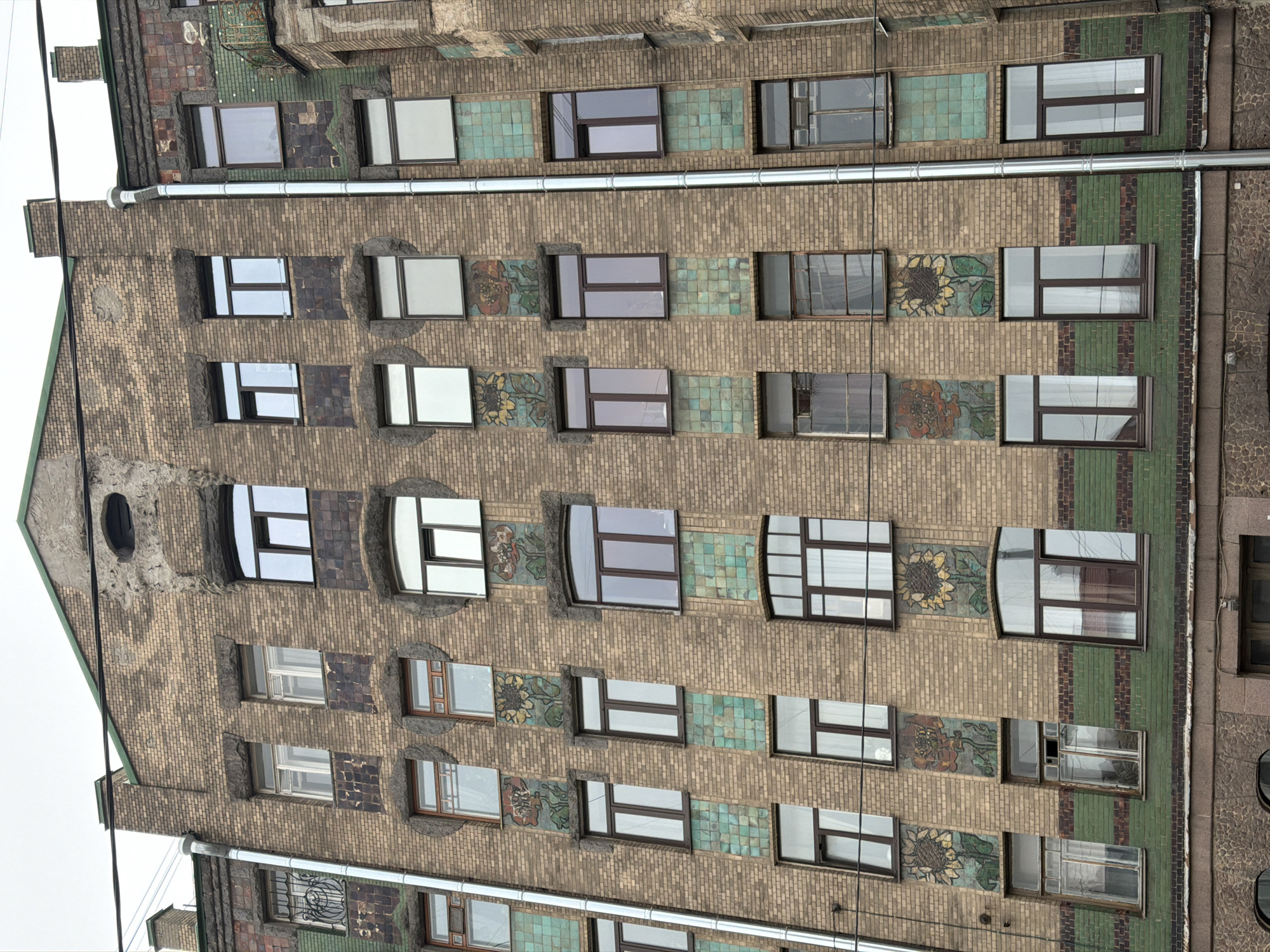 The image shows the facade of a building in Saint Petersburg, Russia. The building is several stories tall, with a brick exterior. It features decorative elements between the rows of windows, including floral motifs and green tiles. The windows are rectangular, some with multiple panes. A metal pipe runs horizontally along the facade. The building's architectural style is unique and distinctive. The overall scene suggests an old, well-maintained building. The sky in the background is overcast.