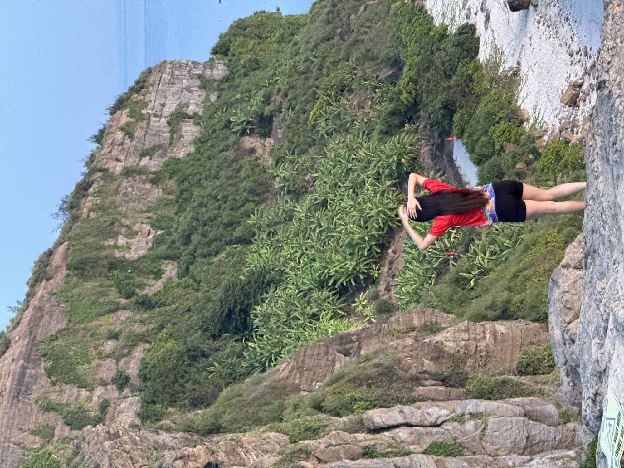 A young woman with long, dark hair, wearing a red t-shirt with colorful accents and black shorts, stands on a rocky cliff overlooking the ocean in Lý Sơn Special Administrative Region, Vietnam. Her hands are placed on the back of her head, and she is facing away from the camera. The cliff face is steep and covered in lush green vegetation, including what appear to be banana trees. The ocean is visible in the background, with a clear blue sky above. A small, sandy beach is situated at the base of the cliff. A red cone marker is visible on the beach near a small body of water.