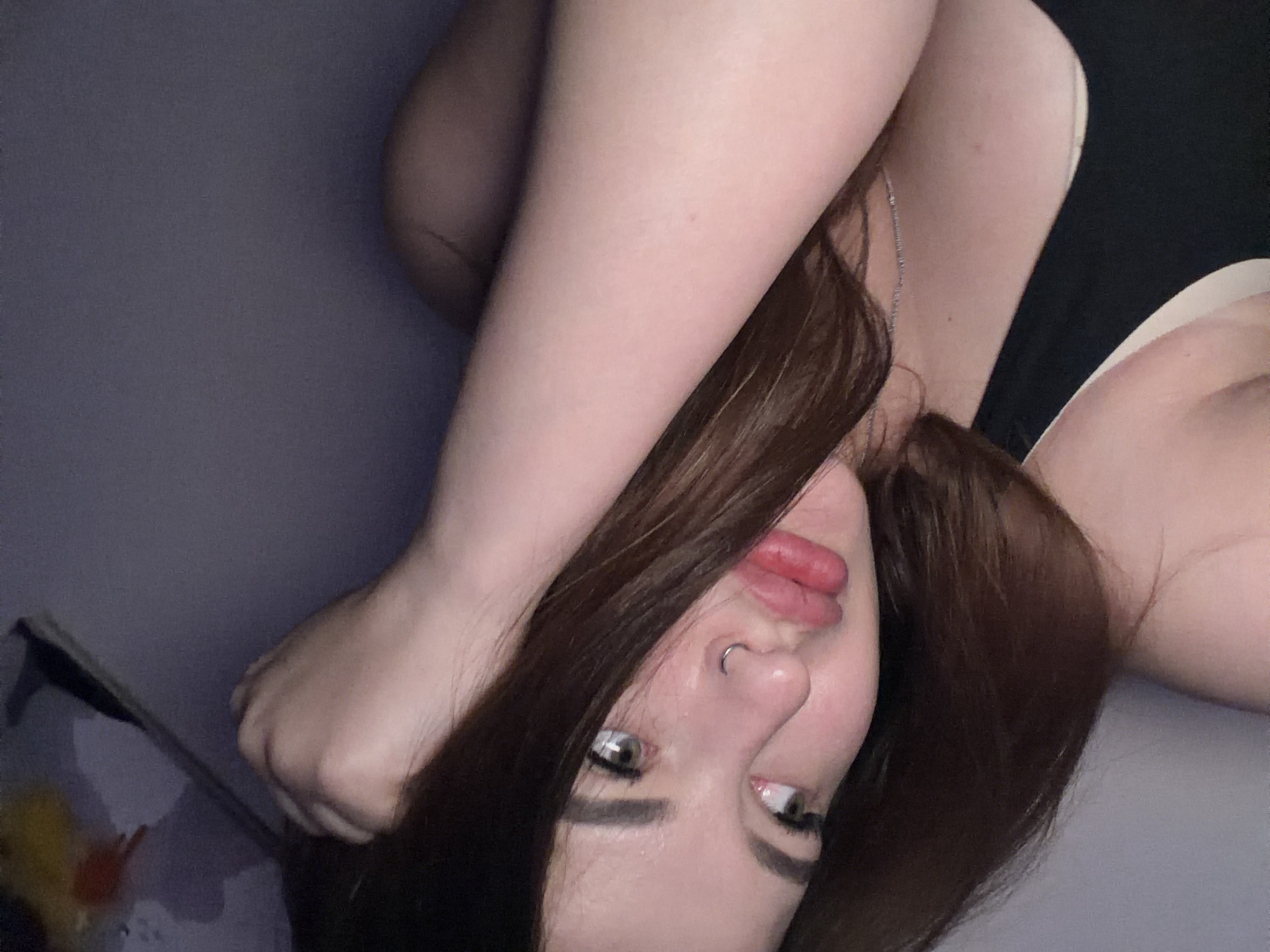 A person is lying on a purple surface. The person has long, brown hair. The person's arms are crossed over their head. The person is wearing a black tank top and a light-colored bra. A silver necklace is visible on the person's chest. The person has a nose ring. The person's eyes are green. The location is not in the city of Krasnodar, Russia.