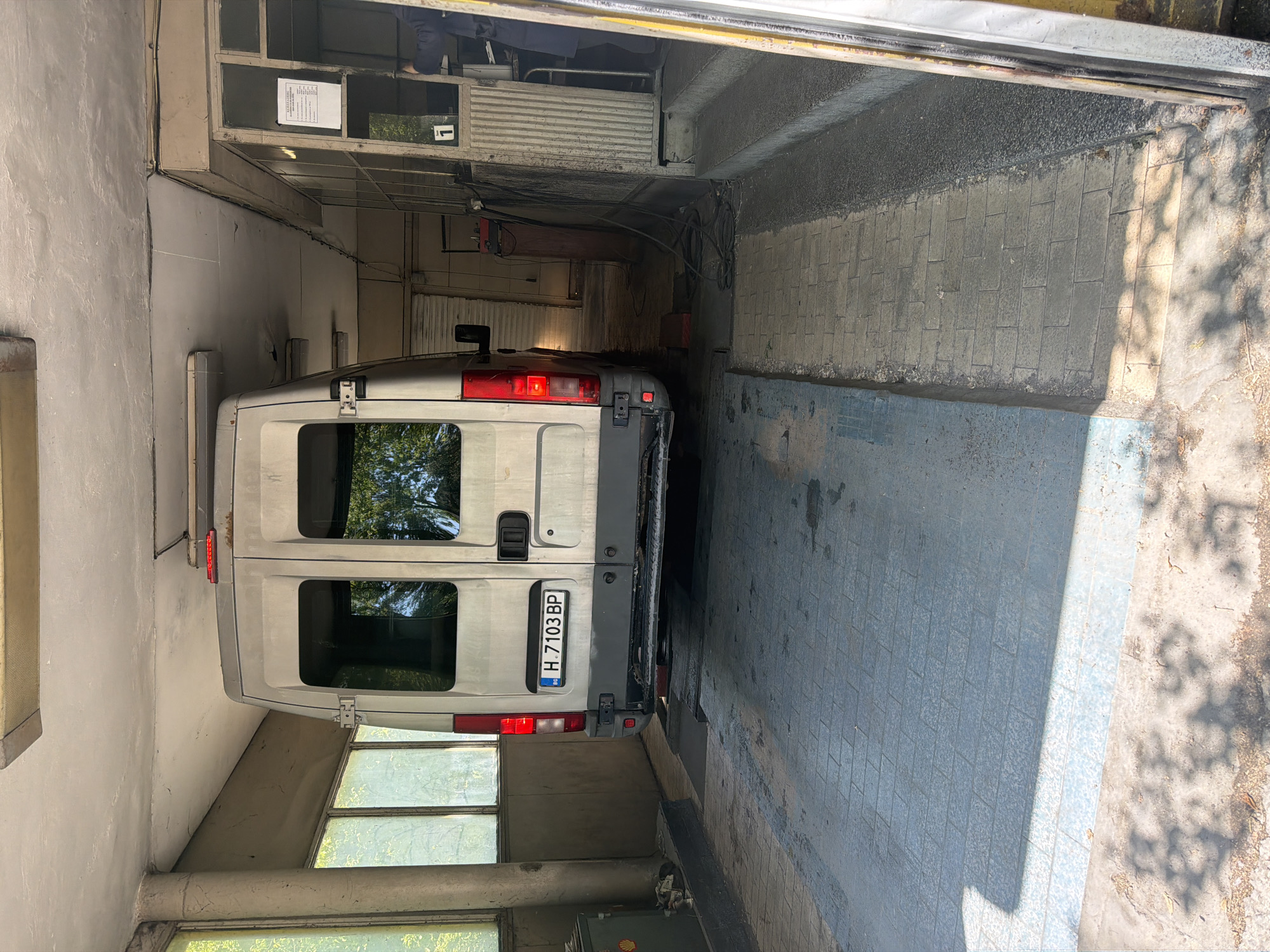 A light-colored van is reversed into a service bay or car wash station, viewed from its rear. The van, possibly a Fiat Ducato or similar model, has two rear doors with windows, and its taillights are visible. A Bulgarian license plate, "H 7103 BP," is centrally mounted on the rear.
The bay features tiled flooring, with a section of blue-grey tiles closer to the van and lighter-toned, possibly worn, tiles further out, leading to rough concrete or asphalt beyond. The walls are made of concrete or cinder blocks, with visible utility elements like pipes, electrical conduits, and cables running along them. Above the van, various machinery components, including what appears to be part of an automated washing system or an overhead door mechanism, are visible.
Bright daylight from outside casts strong shadows across the tiled ground and illuminates the interior, suggesting a sunny daytime. Light also reflects through the van's rear windows, showing reflections of green foliage outside.
In the upper left area of the bay (visible in cropped sections), there is a small elevated office or control area with a shelf holding a printed document, though the text is illegible. A person's legs and feet are partially visible in this area. A number "1" is also visible on a surface in this vicinity. Near the van's rear on the ground, a container with a partial "Shell" logo can be seen. The general appearance of the facility suggests it is well-used.