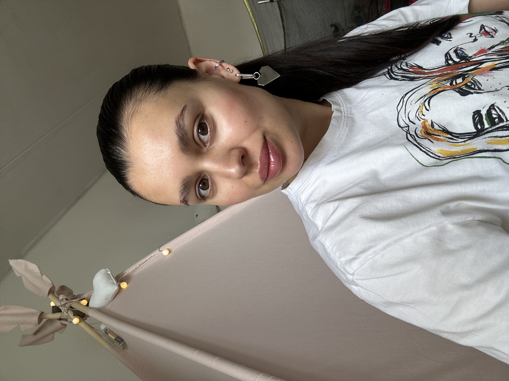 The image is an angled, close-up self-portrait of a young woman indoors in Chaikovsky, Russia. The woman has dark hair pulled back from her face and looks directly at the camera with a gentle, slightly smiling expression. She has brown eyes, full glossy lips, and a natural flush on her cheeks. She is wearing a white t-shirt adorned with a colorful, abstract line-art graphic depicting stylized faces.
Her right ear is visible, showcasing multiple piercings, including small hoops and studs, and a prominent silver-toned earring with a heart-shaped dangle. Her long, dark hair extends over her shoulder and across her shirt.
The background indicates an indoor setting, featuring a light-colored fabric structure, possibly a teepee or canopy, supported by wooden poles. Warm-toned fairy lights are strung around these poles, with a cylindrical battery attached, likely powering the lights. A small white fabric object is also visible near the lights. The scene suggests a cozy and decorated personal space. There is no visible text in the image.