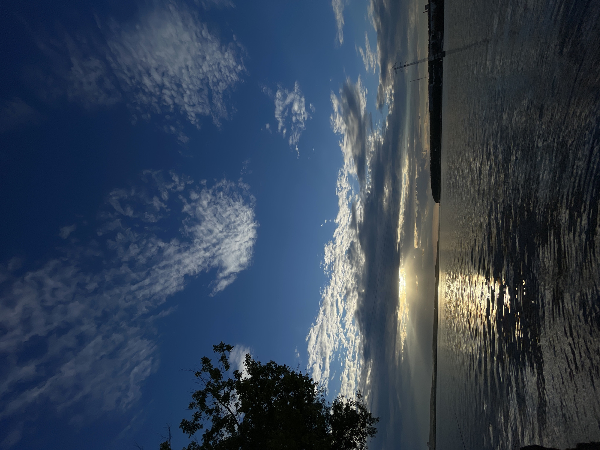 The image depicts a serene body of water under a dynamic sky. The sun, partially obscured by clouds, casts a golden glow and creates a shimmering reflection on the water's surface. Wispy clouds streak across the deep blue sky, suggesting a transition in weather or time of day. On the left side of the frame, the silhouette of a tree branches out, adding a natural element to the scene. In the distance, a shoreline is visible, with landmasses and some man-made structures, including what appears to be a tall transmission tower near the right edge of the frame. The overall impression is one of natural beauty and tranquility.