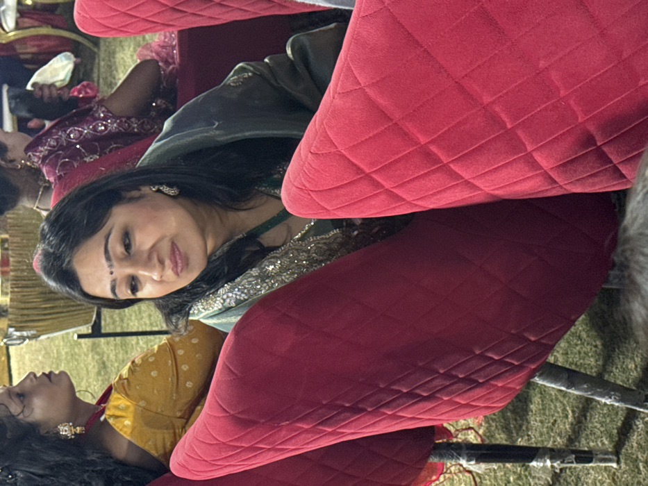 A woman wearing an ornate green and gold dress sits among other people. She has dark hair and is wearing earrings. The surrounding attendees are adorned in similar colorful garments. Red chairs with a quilted pattern fill the scene, obscuring portions of the subjects. The photo appears to have been taken outdoors.