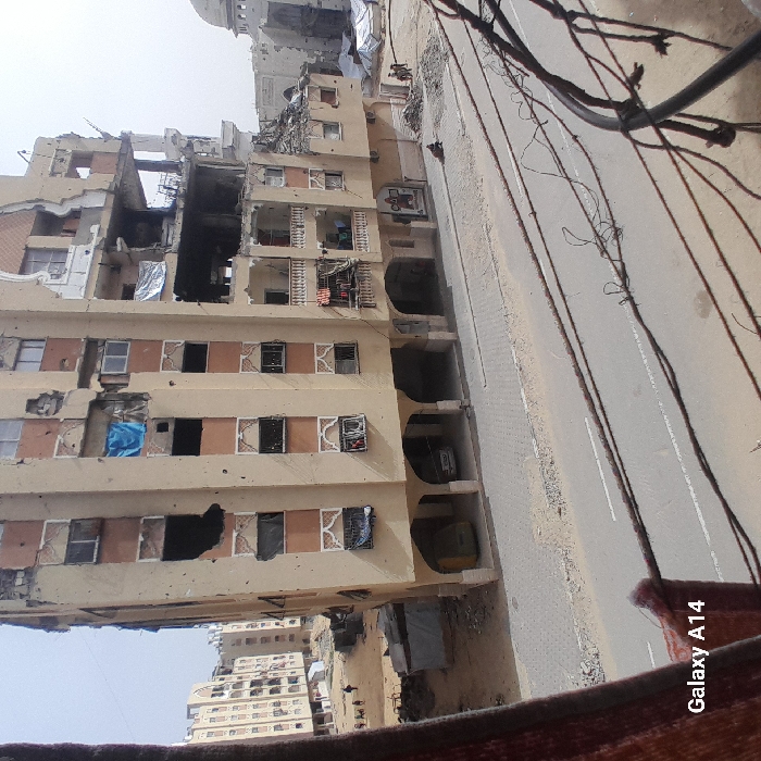 The image depicts a devastated urban landscape in Khan Yunis, Palestinian Territory. A prominent, multi-story, beige-colored residential building occupies the central frame, exhibiting significant structural damage including large gaping holes, exposed interiors, and numerous smaller impact marks on its facade. Some windows are missing panes, and various openings are covered with blue or silver tarps. Clothes are visibly hanging on balconies. The ground floor features an arcade with arched entrances, under which a yellow van and a white car are parked.
In the immediate foreground, electrical or communication wires are visible, partially obscuring the view. Below these wires, a paved road with white markings spans the width of the frame, bordered by a grey paver sidewalk.
To the right of the central building, a substantial amount of rubble and dirt is piled. A single individual is seen seated or crouching near this debris, while a small group of approximately three individuals stands further back. In the distant background on the right, a damaged structure with a prominent dome, likely a mosque, is visible amidst further widespread destruction and temporary tarp shelters.
To the left of the central building, more multi-story residential buildings, also appearing damaged, extend into the background. A small group of approximately three to four individuals is visible walking in this area. The overall environment suggests a scene of widespread destruction and displacement under a clear, pale blue sky.
