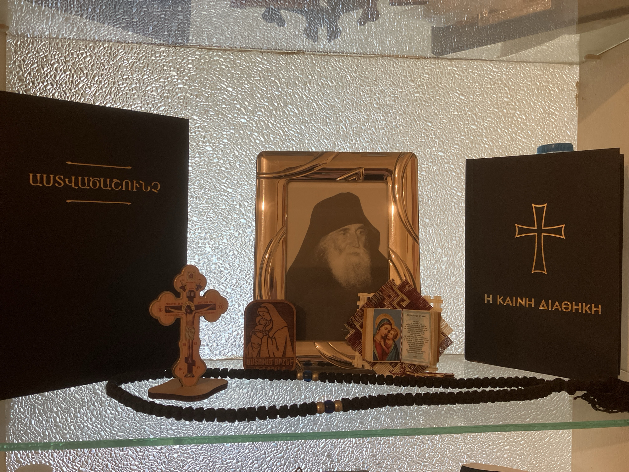 This image displays a collection of religious items arranged on a clear glass shelf, set against a textured, possibly frosted or patterned glass background. The scene suggests a devotional display, likely within a home or sacred space.
On the left, there is a black book with gold Armenian text that reads "ԱՍՏՎԱԾԱՇՈՒՆՉ", which translates to "Bible". To its right, centrally placed, is a gold-framed black and white photograph depicting an elderly bearded monk wearing a dark habit, gazing directly forward. Further to the right, another black book features a gold cross symbol and Greek text reading "Η ΚΑΙΝΗ ΔΙΑΘΗΚΗ", meaning "The New Testament".
In the foreground, a black knotted prayer rope, possibly a komboskini or chotki, with a few blue beads, is draped across the shelf, extending from the left to the right. Resting on the prayer rope, in front of the books and photograph, are several smaller devotional objects. These include a wooden crucifix with intricate carvings of Jesus on the cross and text such as "IC XC" (Jesus Christ) and "NIKA" (Victory), along with other Cyrillic or Greek inscriptions. Next to the crucifix is a small wooden plaque carved with an image of the Virgin Mary holding the infant Jesus, accompanied by Armenian text "ԱՍՏՈՒԾՈ ՈՐԴԻՆ" (Son of God). Behind these, a small miniature icon or print of Mary and child rests on a piece of decorative patterned fabric, with faint, illegible text beside it.
The lighting appears soft, highlighting the textures of the background and the metallic gleam of the photo frame. The combination of Armenian and Greek texts and Orthodox Christian iconography points to a context within Eastern Christianity. No activity is taking place; it is a static display of revered objects.