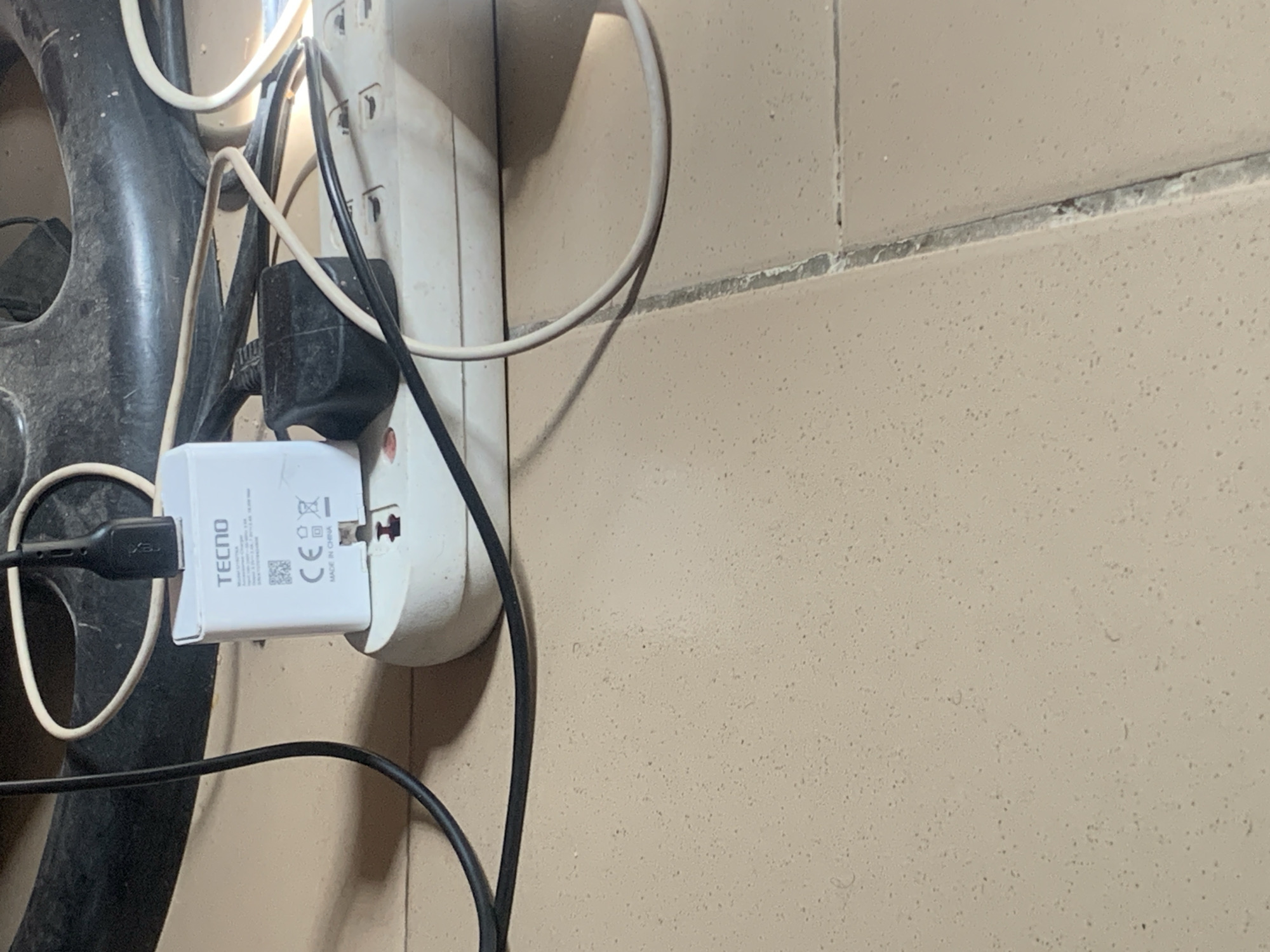 The image depicts a close-up, low-angle shot of a power strip with several plugs and cords connected. A white TECNO brand phone charger is plugged into one socket of the power strip. A black USB cable extends from the TECNO charger, appearing to be connected to another device or power source that is not fully visible. Another black plug, possibly for a laptop or other electronic device, is also plugged into the power strip, with a beige cord extending from it. The power strip itself is white and positioned on a tiled floor. The floor tiles are light beige with darker speckles. In the background, a dark, circular object, possibly a fan or a part of a larger appliance, is visible. The overall scene appears to be indoors, likely in a residential or office setting, and does not provide any specific geographical indicators to confirm its location in Jauro Yinu, Nigeria. The lighting suggests an indoor environment with artificial light. There are no people visible in the image.