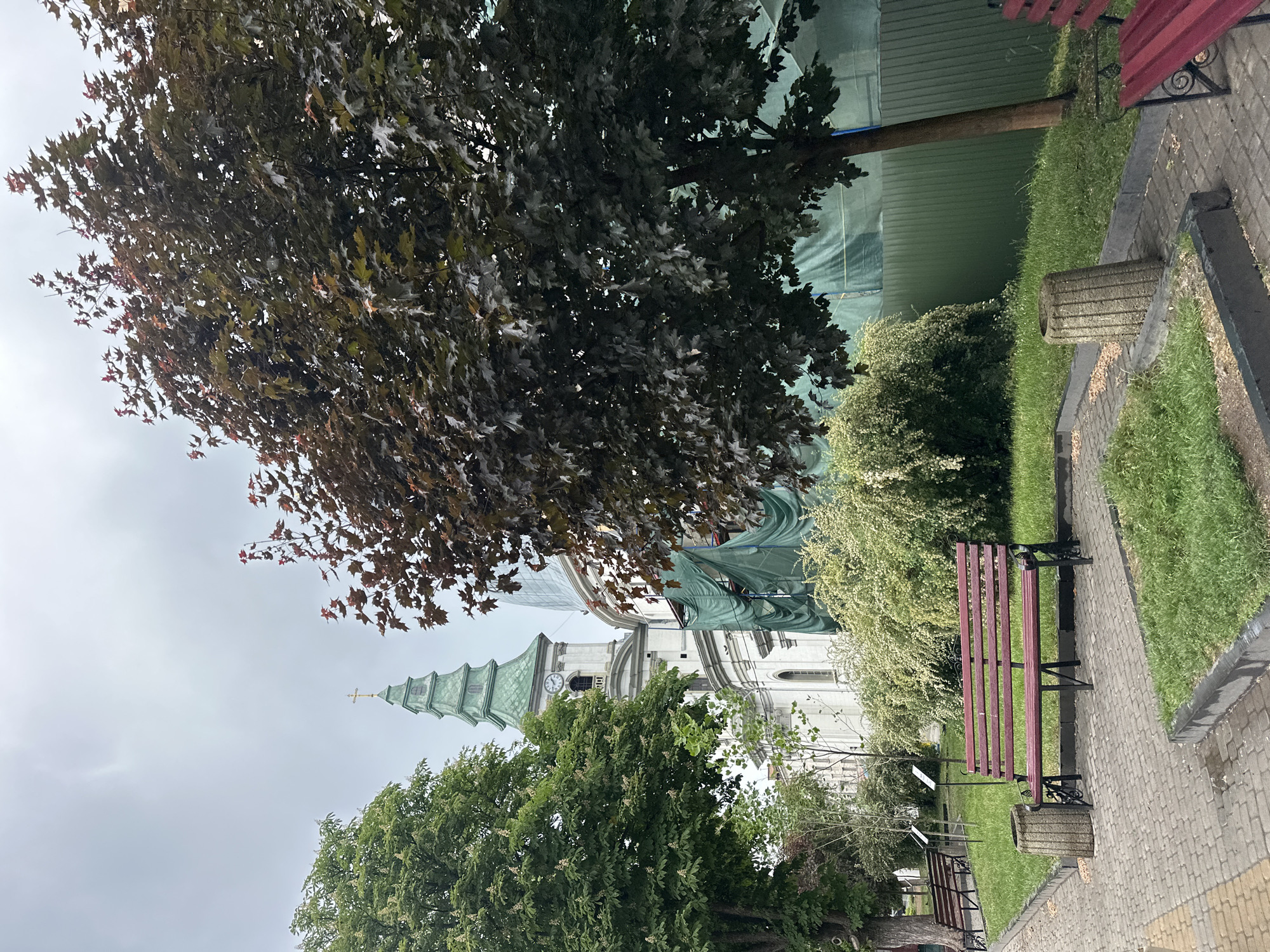The image captures a scene in Ternopil, Ukraine. A church tower with a green spire rises in the background, partially obscured by trees. The building is undergoing maintenance as seen by the tarps covering it. In the foreground, there is a paved walkway with benches alongside grass and shrubbery. A large tree with red leaves dominates the upper left corner of the frame. The sky is overcast.