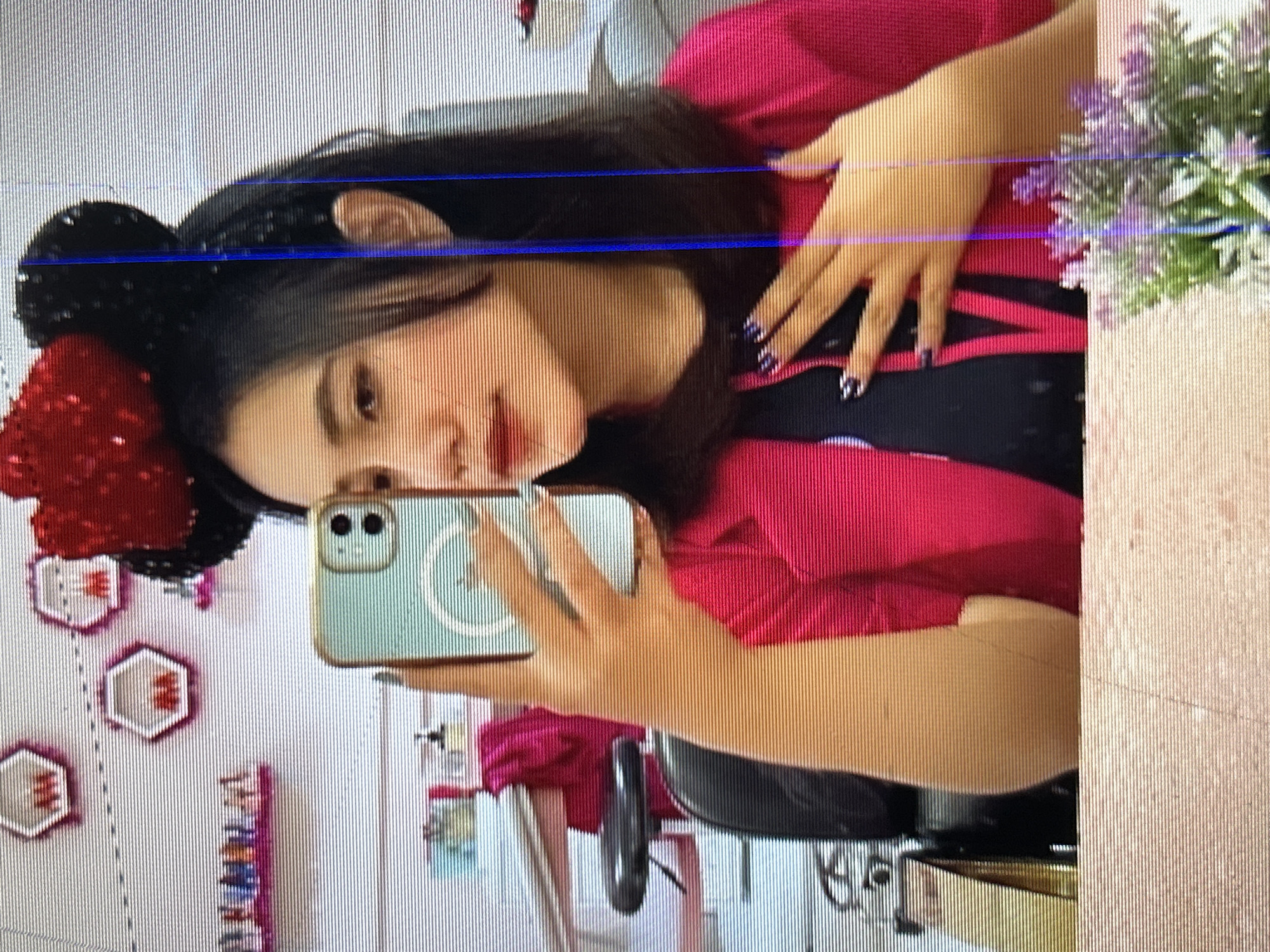 The image is a close-up selfie of a young woman in what appears to be a nail salon or beauty parlor. She is wearing a red sequined Minnie Mouse headband and a bright pink blazer over a dark top. Her right hand is holding up a light blue smartphone, and her left hand is resting on her chest. The background shows shelves on a white wall decorated with hexagonal shelves, some containing small items. There are also glimpses of nail polish bottles and other beauty supplies. The woman's nails are painted with a dark color and intricate designs. The overall impression is of a young woman taking a selfie in a brightly decorated, possibly professional, setting. The specific location in Depok, Indonesia is not discernible from the image alone.