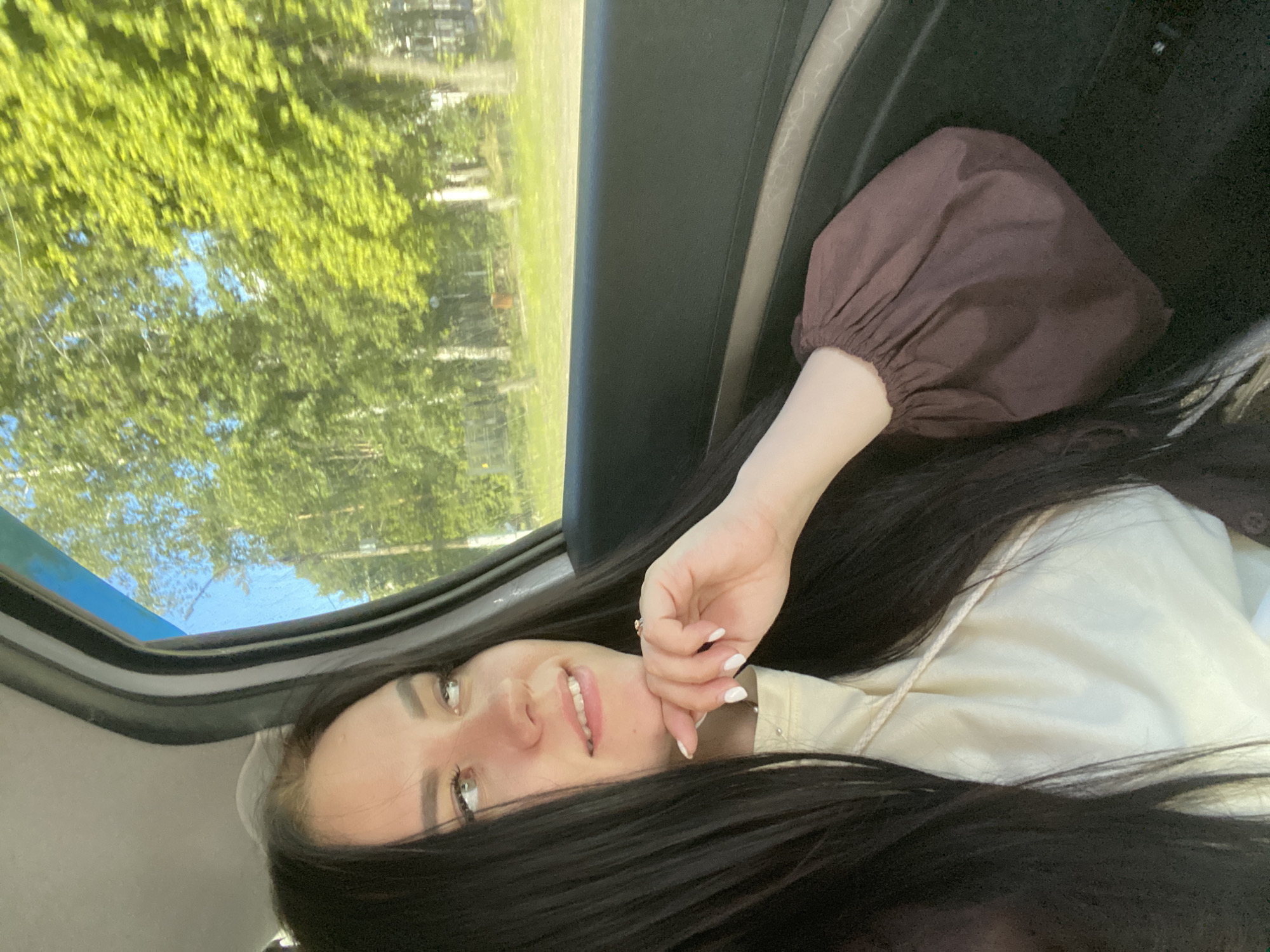 A woman with long, dark hair is lying on her back in a car. She is looking up and smiling slightly. Her right hand is near her face. She is wearing a light-colored top and a brown blouse. Through the car's open sunroof, green trees and a blue sky are visible. The location is possibly in the city of Chaikovsky, Russia, given the request.