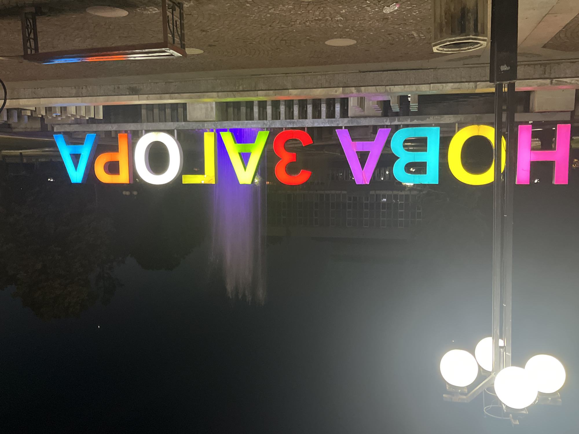 The image depicts a nighttime scene in the city of Nova Zagora, Bulgaria. In the foreground, large, illuminated letters spell out "НОВА ЗАГОРА" in a variety of vibrant colors including pink, yellow, teal, purple, red, green, white, orange, and blue. The letters are mounted on a stone structure in front of a fountain that emits a purple glow. To the left, a streetlamp with four white, spherical lights illuminates the area. Behind the illuminated sign, a building with numerous windows is visible. In the far background, trees and a single bright light in the sky can be seen. A dark-colored trash can is positioned near the streetlamp, and a wooden bench with metal legs is in the lower right portion of the image. The ground is paved with cobblestones, and some litter is present on the ground. The overall atmosphere is dark, with the illuminated elements creating points of focus.