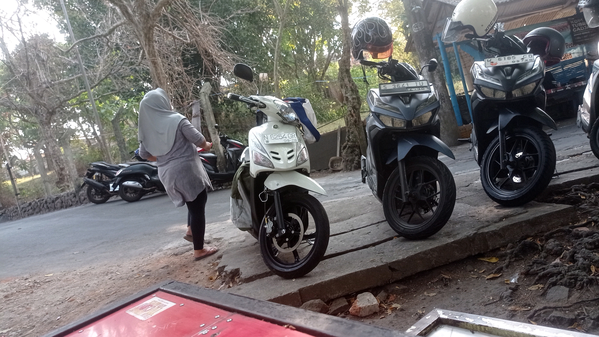 In Mulyoagung, Indonesia, on a roadside lined with trees, a woman wearing a grey hijab and grey tunic over black leggings stands beside a white scooter. The scooter has a license plate that reads "N 6254 GR 09-28". Behind it, parked on the paved road, are several other motorbikes, including two dark-colored scooters with helmets on their handlebars. One helmet is white and branded "XRC", and the other is black with a flame design. The scene appears to be outdoors during daylight, with a natural backdrop of trees and foliage. The foreground shows a red object with metal trim, possibly part of a table or a sign. The ground is a mix of dirt, rocks, and exposed tree roots.
