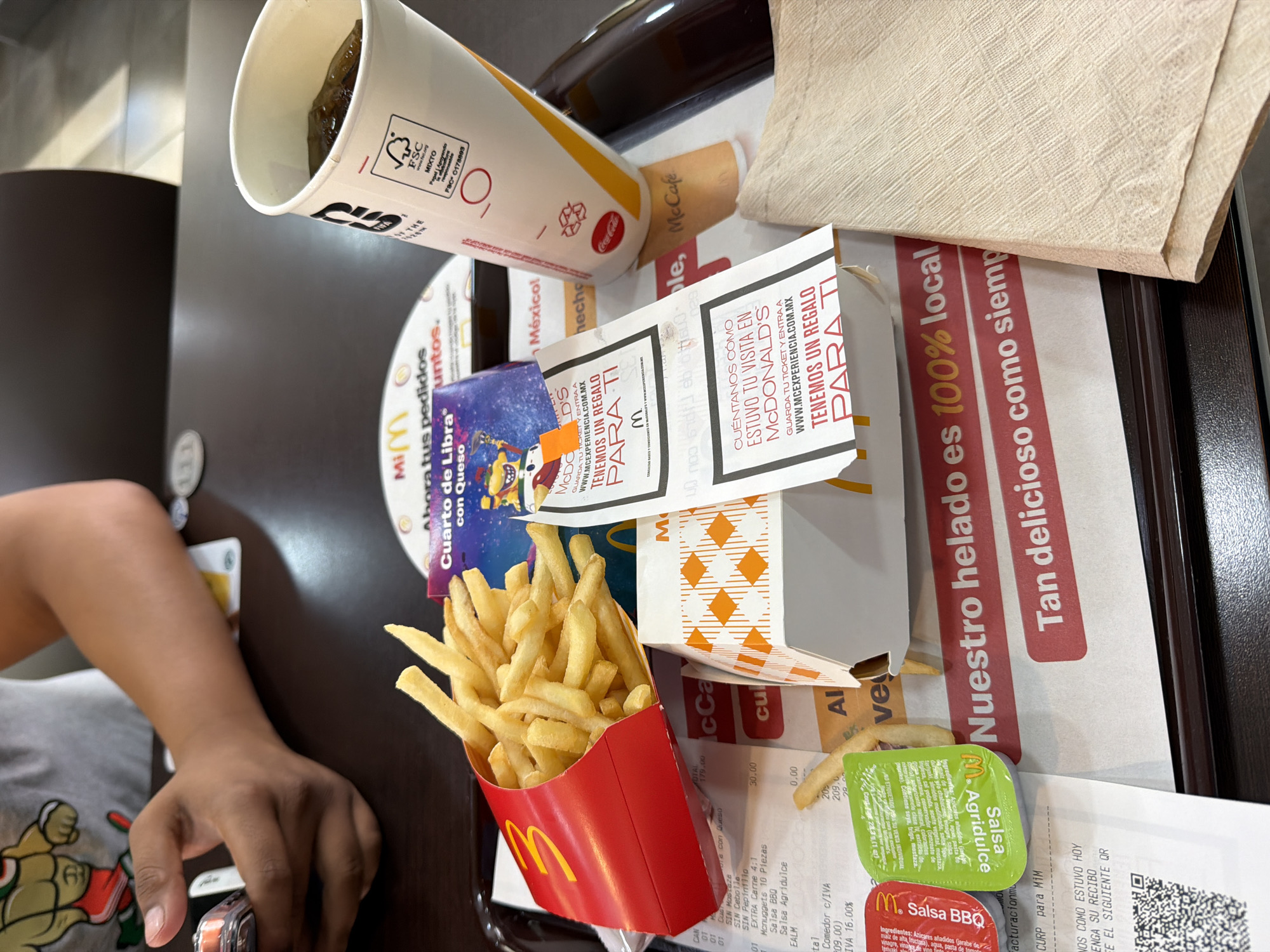 The image shows a table at a McDonald's restaurant, presumably in San Andrés Cholula, Mexico, given the text in Spanish. A person's arm is visible, wearing a gray shirt with a cartoon character on it. The table is covered with McDonald's food and packaging, including a cup with a straw, a box of french fries, a McDonald's wrapper, a napkin, two packets of sauce, a receipt, and promotional material. The promotional material has text about a survey for a free gift. The table has a brown surface with a tray holding the items. The overall setting is a fast-food dining area.