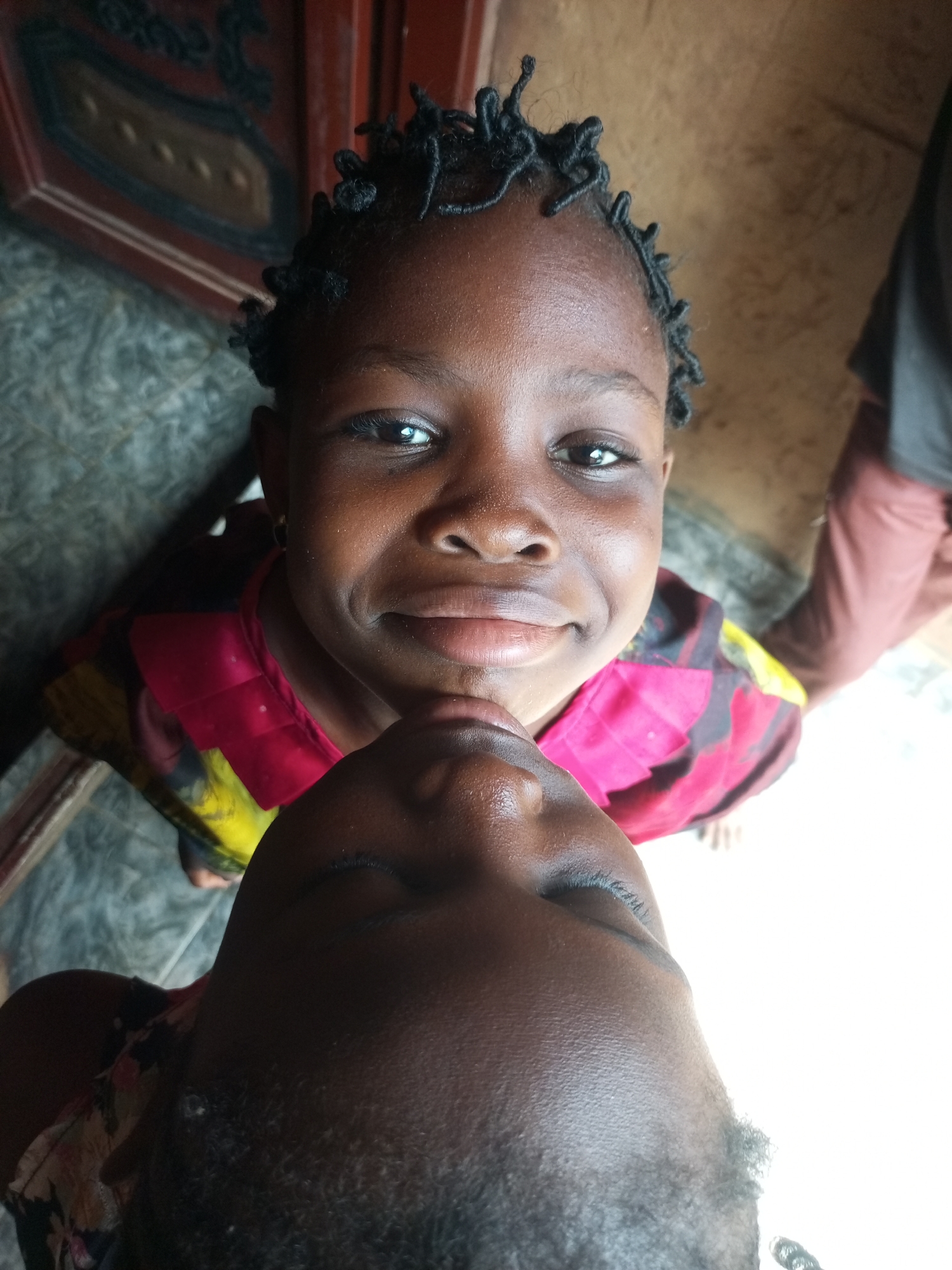 The image shows a close-up, high-angle view of two young Black children. The child on top has dark skin, black braided hair, and is smiling with dimples. They are wearing a brightly colored top with pink and yellow patterns. Their chin rests on the forehead of the child below, who is looking upwards with closed eyes and a slightly parted mouth. The child below has dark skin and is wearing a patterned shirt with floral motifs. The background includes a textured grey floor, a brown wooden surface with intricate dark detailing, and a beige wall. A person's legs and lower torso, wearing light brown pants, are visible in the upper right portion of the frame. The scene appears to be indoors, possibly a residential area in Gombe, Nigeria.