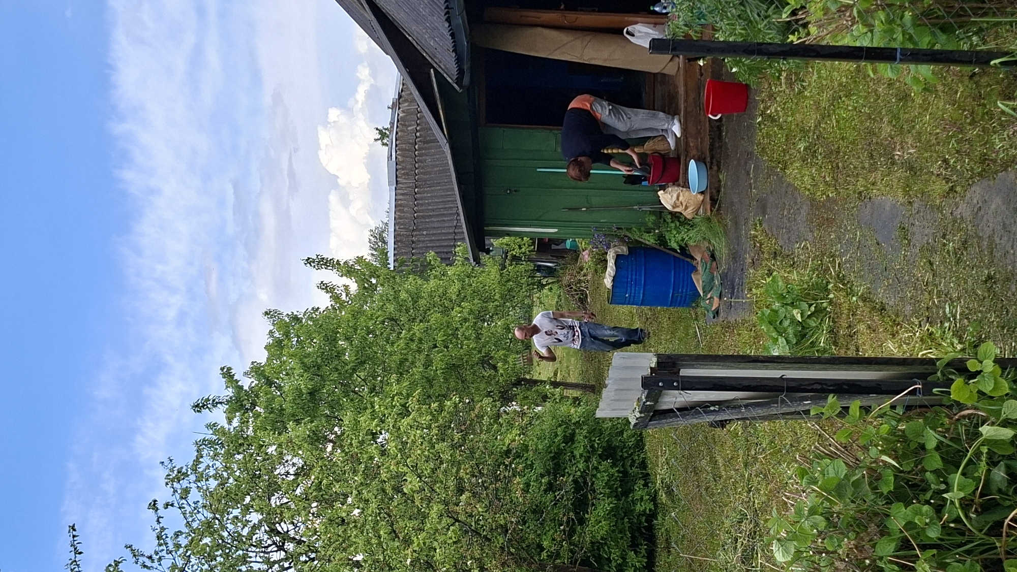 The image shows an outdoor scene in Сваткаўскі сельскі Савет, Belarus. A green wooden structure with a dark gray roof is partially visible. Two people are present. One is bent over, possibly cleaning with a bucket and pole, while the other stands near a tree wearing a white t-shirt and jeans. A blue barrel sits nearby. A pathway leads from the structure through grass and foliage. A partly disassembled wooden structure lies on the ground, close to foliage and a chain link fence. The sky is partly cloudy.