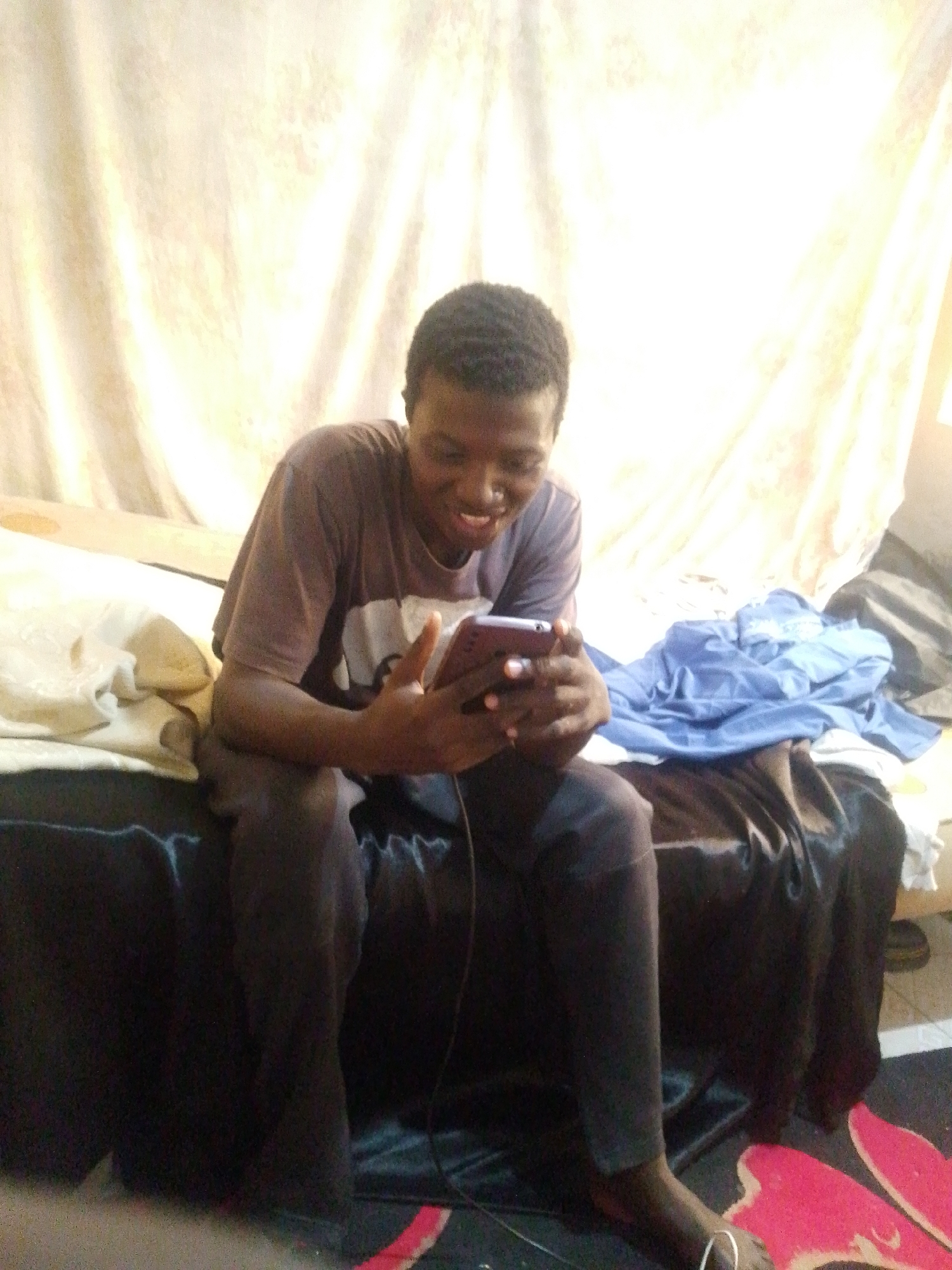 A young Black male with short, dark hair and a warm smile is sitting on the edge of a bed, looking down at a smartphone he holds in his hands. He is wearing a dark t-shirt with a lighter colored graphic on it, and dark pants. The bed is covered with a dark, possibly satin-like fabric, and there are some piles of clothing on the bed behind him, including a blue shirt. A pale, textured curtain hangs in the background. The overall lighting is bright, with a slight overexposure effect. This scene appears to be indoors, possibly in a bedroom, in Jos, Nigeria.