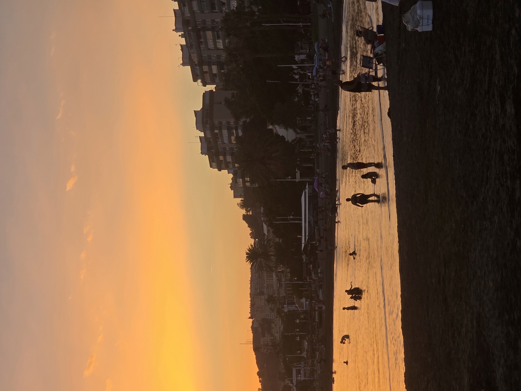 The image depicts a sunset scene at a beach in Málaga, Spain. Silhouettes of people are visible wading in the calm sea and standing on the dark sand. The sky is a vibrant gradient of orange and yellow, with scattered clouds. In the background, a city skyline with apartment buildings and palm trees is visible, illuminated by the warm light of the setting sun. The beach appears to be a popular spot, with many individuals enjoying the evening. The overall atmosphere is serene and picturesque, capturing the beauty of a coastal sunset in Málaga.
