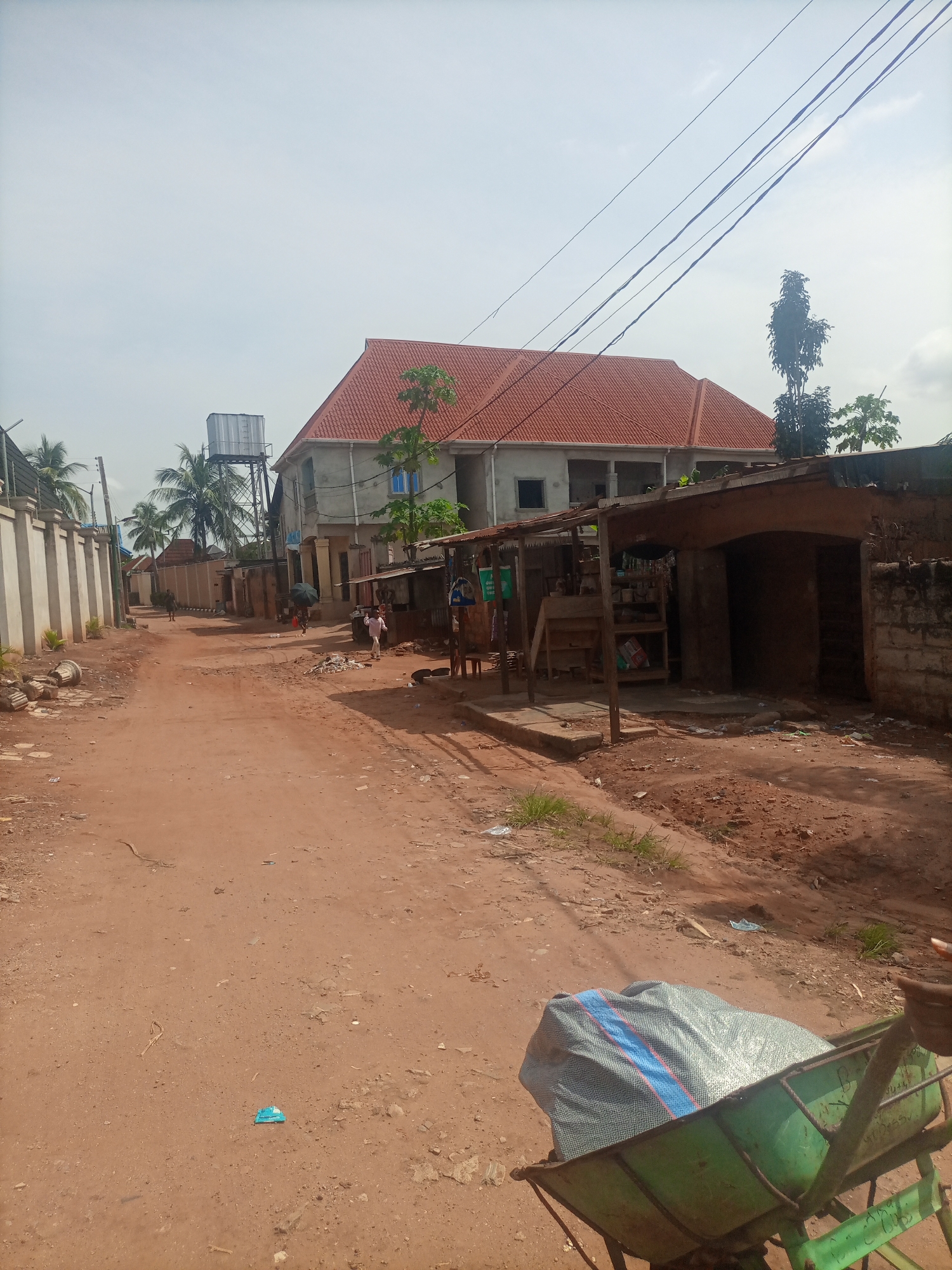 A dirt road in Nkpor, Nigeria, is lined with buildings and vegetation. The road is unpaved and appears to be a main thoroughfare in the area. On the left side of the road, a tall, white concrete wall with vertical lines separates the street from a property with palm trees. Further down the road on the left, a large, two-story building with a red-tiled roof is visible, along with a water tower. On the right side of the road, there are several small shops and stalls with corrugated metal roofs and open fronts. One of these stalls has shelves displaying various items. A person is seen walking in the distance on the left side of the road. In the foreground on the right, a green wheelbarrow filled with a large, grey, striped bag is present. The sky is overcast, and the overall scene suggests a typical day in a Nigerian town with a mix of residential and commercial structures.