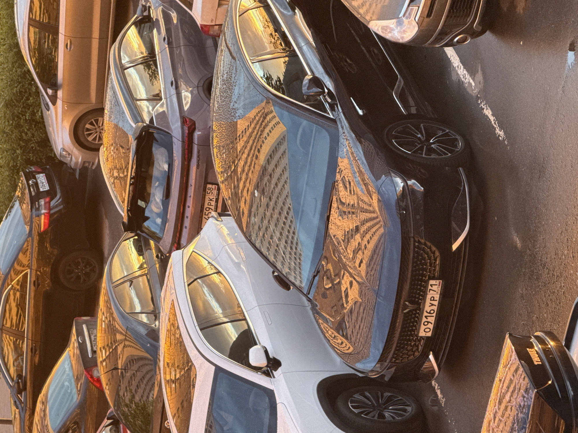 This media file depicts a vertical, eye-level shot of several parked cars on a paved surface, likely a street or parking lot, in Drozhzhino, Russia. The scene is bathed in the warm, golden light of either sunrise or sunset, casting long shadows and creating strong reflections on the cars' surfaces. The sky appears clear, with no visible clouds.
The primary subjects are the cars themselves. A silver SUV is parked on the left, partially obscured by grass. Next to it are several darker-colored cars, including a black sedan with a prominent front grille and a license plate reading "0916 YP 71 RUS". The reflections on the cars show the facade of a multi-story building, suggesting an urban or suburban environment. Another white vehicle is parked to the right, also reflecting the building. A dark-colored car with a "THULE" logo visible on its rear is also present.
The activity taking place is simple: the cars are parked. There is no human presence or overt action. The notable details include the strong directional light, the reflections on the cars, and the Russian license plate, which provides a specific location clue. The overall mood is tranquil, perhaps even serene, due to the peaceful setting and the quality of the light. The image captures a moment of stillness in an everyday urban environment.