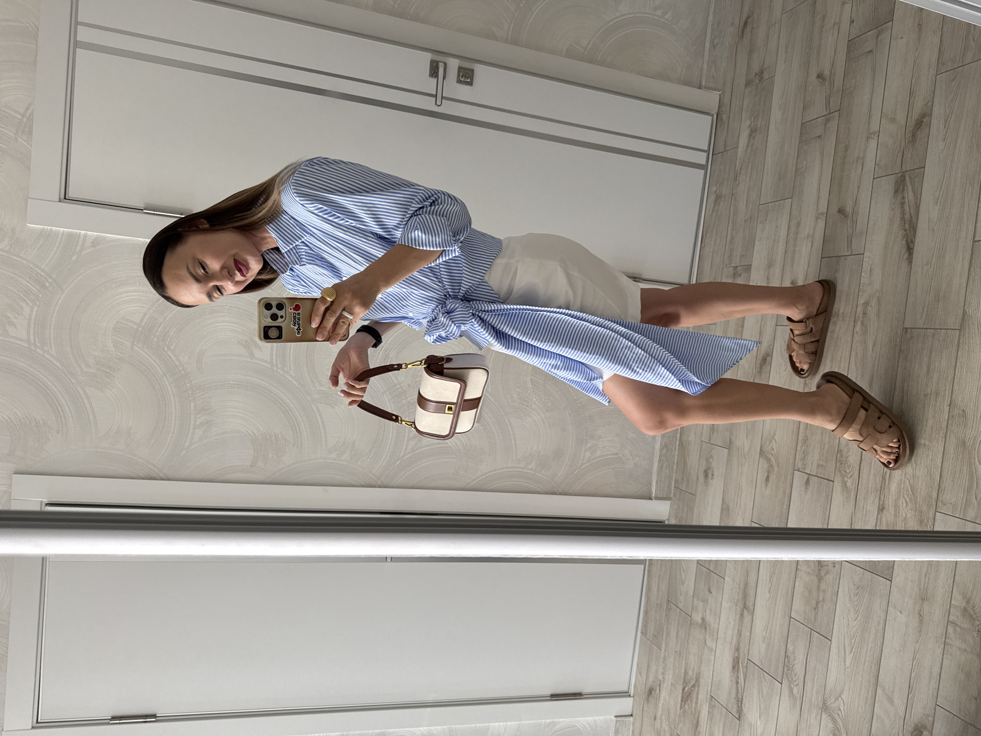 A woman stands in front of a mirror, taking a selfie. She is wearing a blue and white striped shirt dress, a white skirt, and brown sandals. She is also holding a small, cream-colored handbag with brown leather trim. The room appears to be a bedroom or dressing room, with a wooden floor and textured wallpaper. The lighting suggests it is daytime. The woman has her hair down and is wearing makeup, including lipstick. She is smiling subtly. The image is taken from a low angle, and the woman's reflection is visible in the mirror.