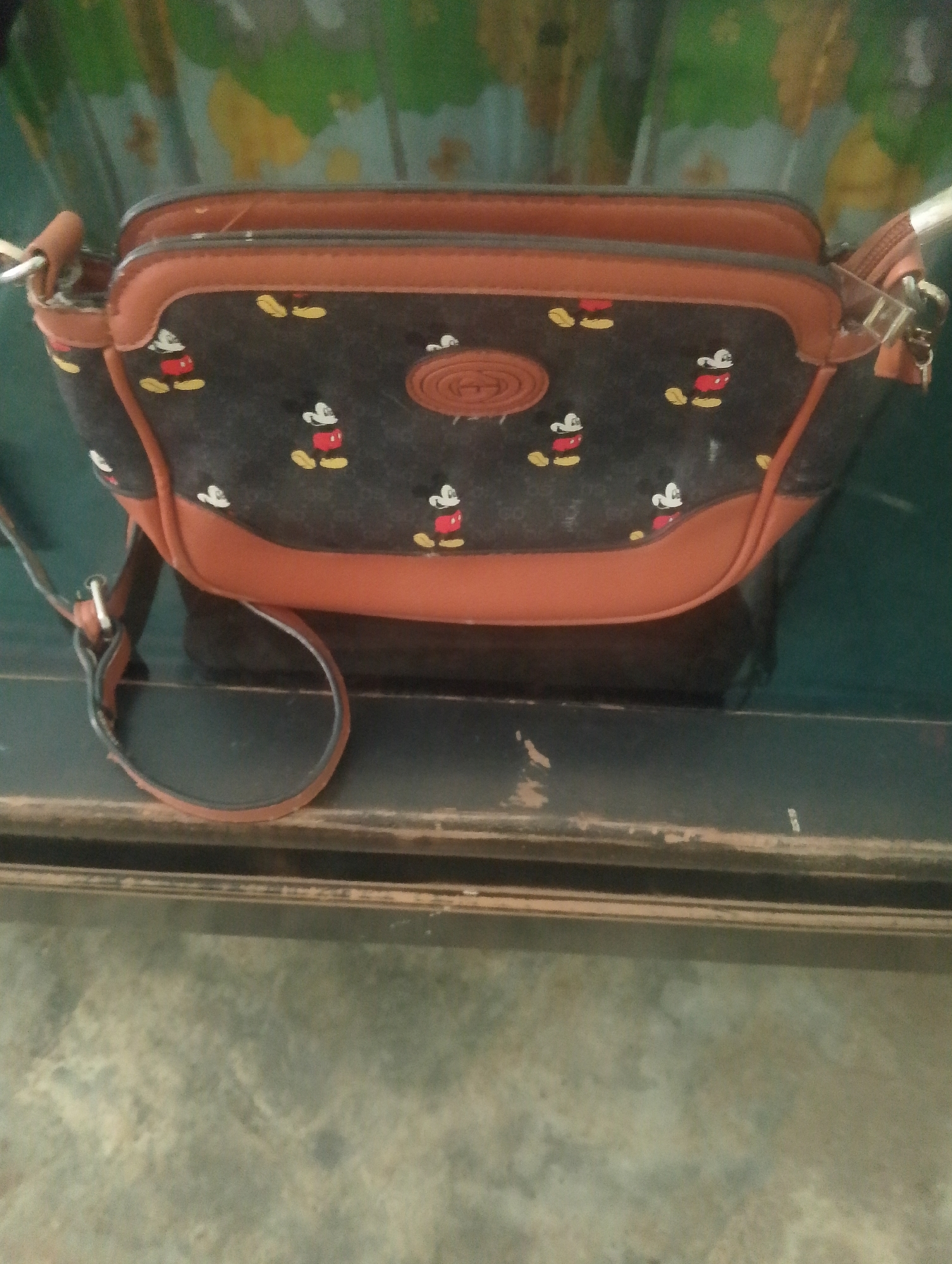 A dark brown and tan handbag with a Mickey Mouse pattern is resting on a dark, reflective surface. The handbag has a circular emblem on the front. The background appears to be a wall with a green and yellow pattern. The image is undated and the location is unknown.