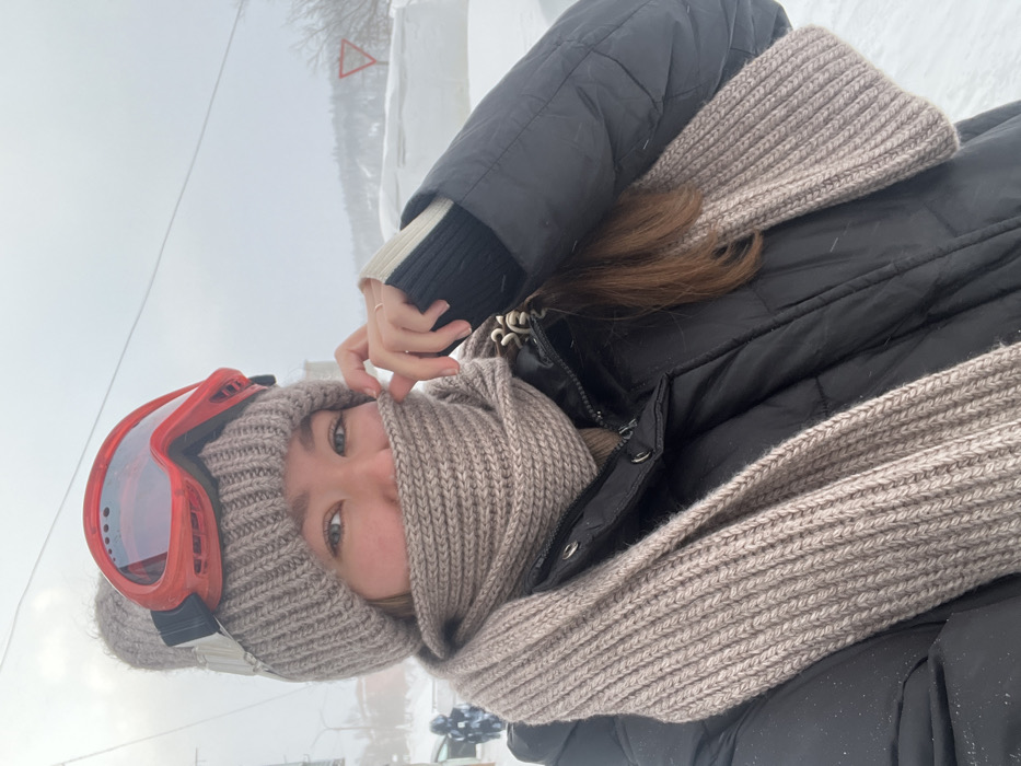 A person wearing ski goggles, a beanie, and a scarf poses in a winter setting. They are also wearing a black puffer jacket and a striped sweater. Their hair is brown and their eyes are visible. The background shows a snowy landscape with possible signage. The photo appears to be taken at a tilted angle.