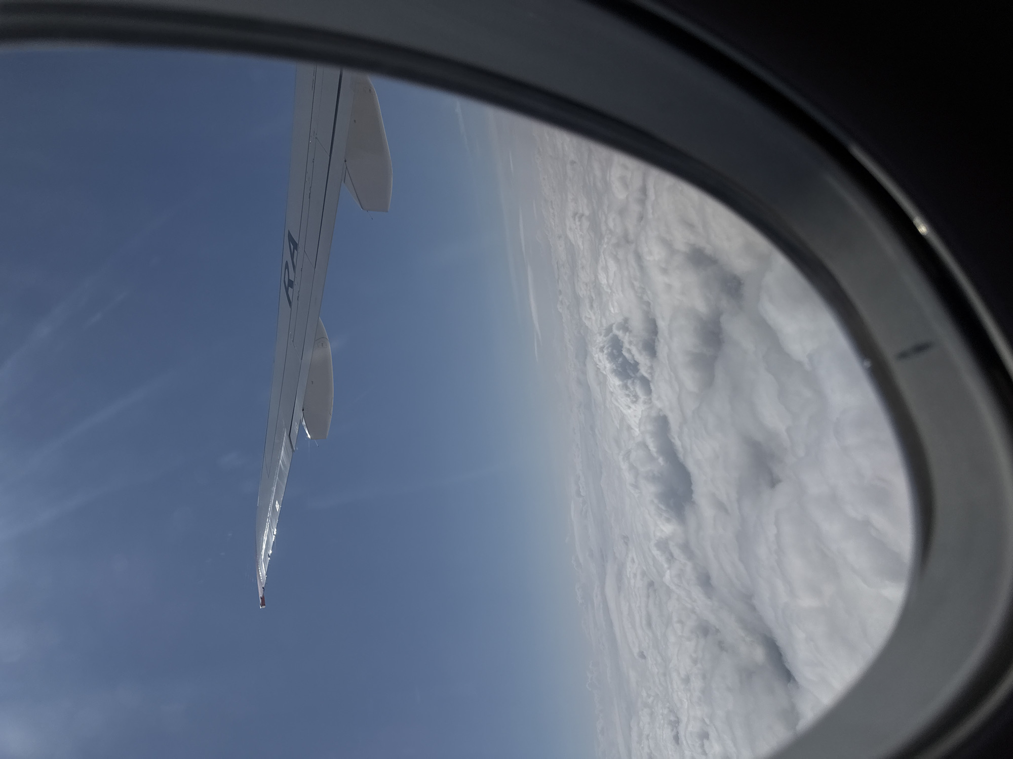 The image is a view from an airplane window, looking out at the sky and clouds. An airplane wing is visible in the upper portion of the frame, with the letters "RA" and some other text on it. Below the wing, a vast expanse of white clouds stretches to the horizon under a bright blue sky. The window frame is dark and partially obstructs the view. The image does not contain any information or visual cues that could place it in the city of Staroe Mart'yanovo, Russia, or any other specific location.