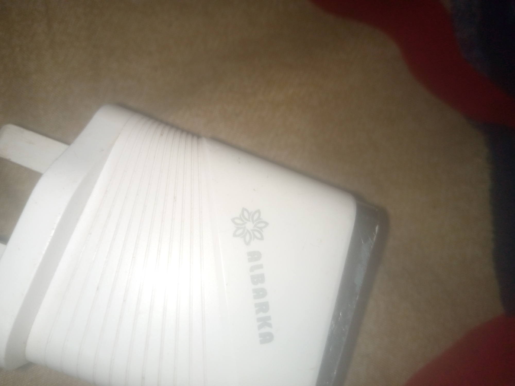 A white wall charger with a logo and text reads "ALBARKA". The charger is on a textured beige surface.