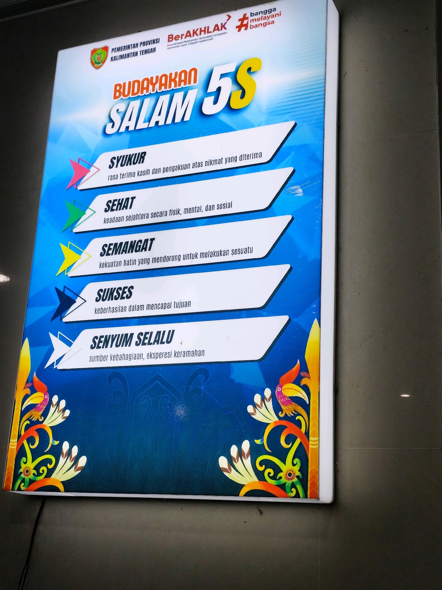 The image shows a brightly lit, rectangular sign mounted on a dark tiled wall. The sign displays the slogan "BUDAYAKAN SALAM 5S" in large, bold lettering, with "BUDAYAKAN" and "SALAM" in orange and "5S" in yellow. Below this, five principles are listed in white boxes with colored arrows pointing to them: SYUKUR (gratitude), SEHAT (healthy), SEMANGAT (enthusiastic), SUKSES (successful), and SENYUM SELALU (always smile). Each principle has a brief definition underneath. The sign features decorative elements of colorful birds and floral motifs in the corners, set against a blue background with subtle patterned designs. The top of the sign includes logos for "PEMERINTAH PROVINSI KALIMANTAN TENGAH" and "BerAKHLAK" with the slogan "bangga melayani bangsa". The location is identified as Palangka Raya, Indonesia, though this is not visible in the image itself. The sign is illuminated from within, casting a soft glow. The wall appears to be an indoor setting, possibly a public building or office. There are no people visible in the image.