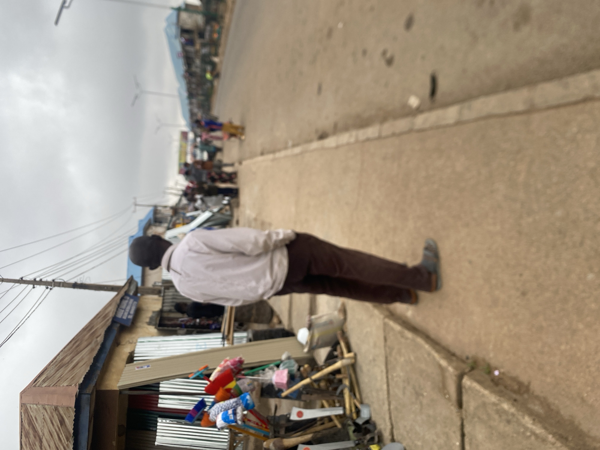 A man stands on a sidewalk in Bukuru, Nigeria. The street is lined with buildings, and people walk in the distance. Debris and merchandise clutter the edge of the sidewalk, next to a building made of corrugated metal. The sky is overcast.
