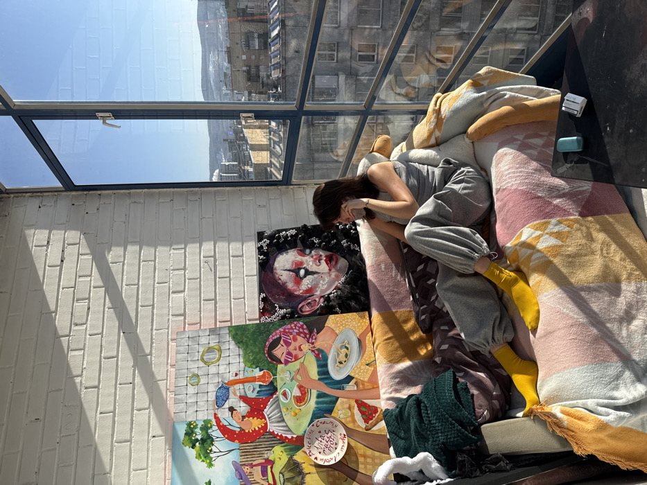 An interior scene depicts a young adult female seated comfortably on a sofa in a room with a large window overlooking the city of Krasnoyarsk, Russia. The specific location within Krasnoyarsk is an elevated position, offering a panorama of multi-story urban buildings, a clear blue sky, and distant snow-capped hills and mountains. A large industrial chimney emitting smoke is visible in the distance.
The female subject has dark hair and wears a light gray tank top, gray sweatpants, and bright yellow socks. She is positioned facing towards the large, dark-framed window, with her left hand near her face. The sofa is draped with a multi-colored patterned blanket featuring geometric shapes in muted tones, and a dark green knitted blanket is on its left armrest.
Behind the sofa, leaning against a white painted brick wall, are two distinct artworks. The left painting is vibrant and colorful, depicting women engaged in leisure activities in a Mediterranean-style setting, featuring lemon trees and a "Dolce Vita" motif. The right painting is darker, portraying a stylized, pale face with red lips and dark eye makeup, resembling a clown or a figure adorned with small white pearl-like objects on a dark background. Strong sunlight enters through the window, casting distinct shadows of the window frames onto the brick wall. On the dark floor in the foreground, a small light blue cylindrical object and a white rectangular object are visible.