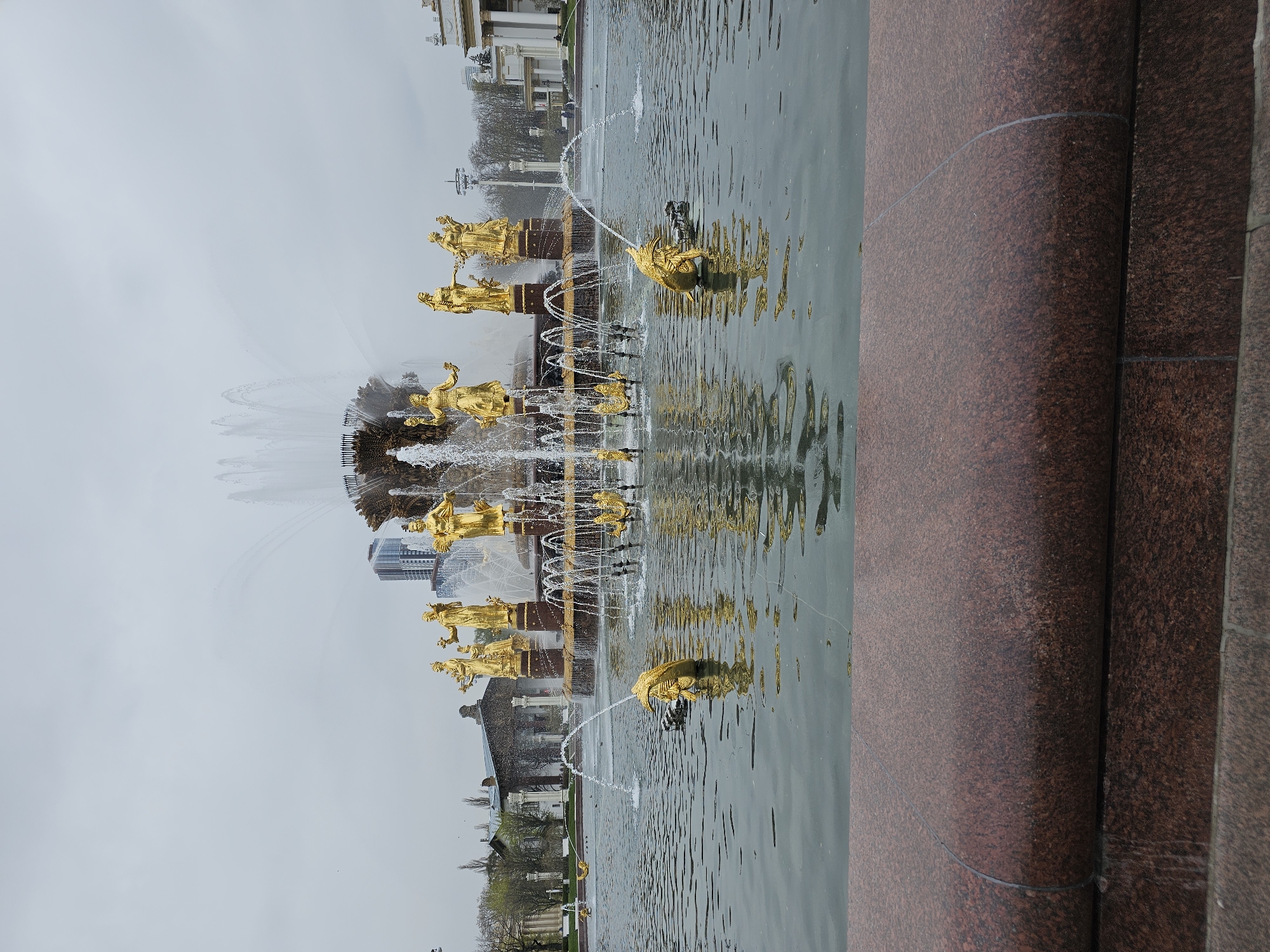 The image shows the Friendship of Nations fountain in Moscow, Russia. The fountain features sixteen gilded statues representing the former Soviet republics. Water cascades from the statues and jets into the pool below, reflecting the golden figures. The foreground includes a portion of the fountain's red granite base. In the background, buildings and trees are visible under a cloudy sky.