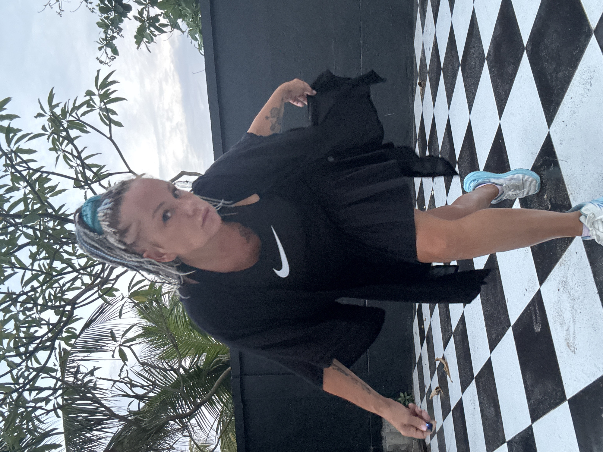 In Kuta, Indonesia, a woman with braided hair, accented with blue streaks, stands on a black and white checkered floor. She is wearing a black Nike t-shirt and a black open-front overlay that extends below her knees, along with a black skirt. Her left hand is extended, holding a small blue object. She is looking towards the camera with a serious expression. Behind her, a dark wall is visible, with lush green trees and foliage framing the top of the image against a cloudy sky. The overall setting appears to be an outdoor area with a modern aesthetic.