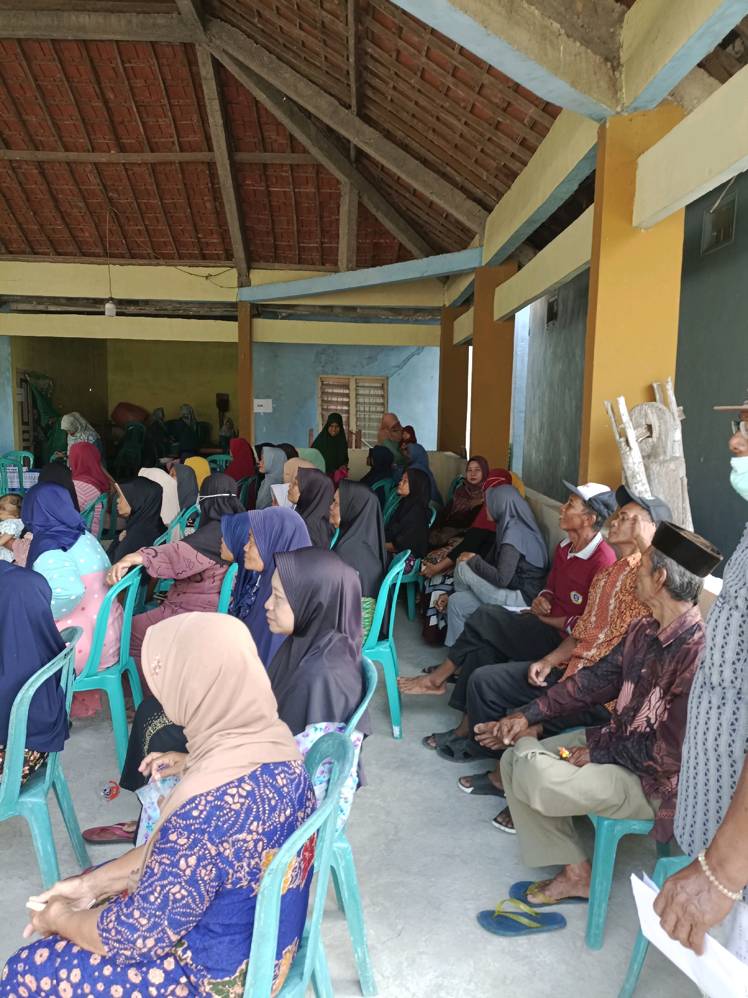 In Rancang Kencono, Indonesia, a group of individuals, primarily women wearing hijabs, are seated on turquoise plastic chairs under a covered outdoor area. The area has a tiled roof supported by wooden beams and columns painted in yellow and blue. The floor is concrete. Some men are also present, seated on chairs and on the floor. Several individuals appear to be listening attentively. In the background, there are doorways leading to interior spaces, and some objects are visible, including a white lamp hanging from the ceiling. The overall scene suggests a gathering, possibly a community meeting or event.