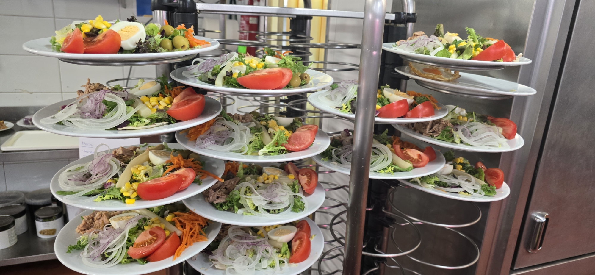 The image shows a commercial kitchen in Sallent de Gállego, Spain, featuring a multi-tiered serving rack filled with individual plates of salads. Each plate contains a salad composed of mixed greens, sliced tomatoes, sliced hard-boiled eggs, corn kernels, olives, and shredded carrots. Some salads also include shredded tuna and thinly sliced red onions. The rack is silver and appears to be made of metal. In the background, there are stainless steel appliances and shelving, typical of a professional kitchen environment. The lighting suggests an indoor setting. There are no people visible in the image.