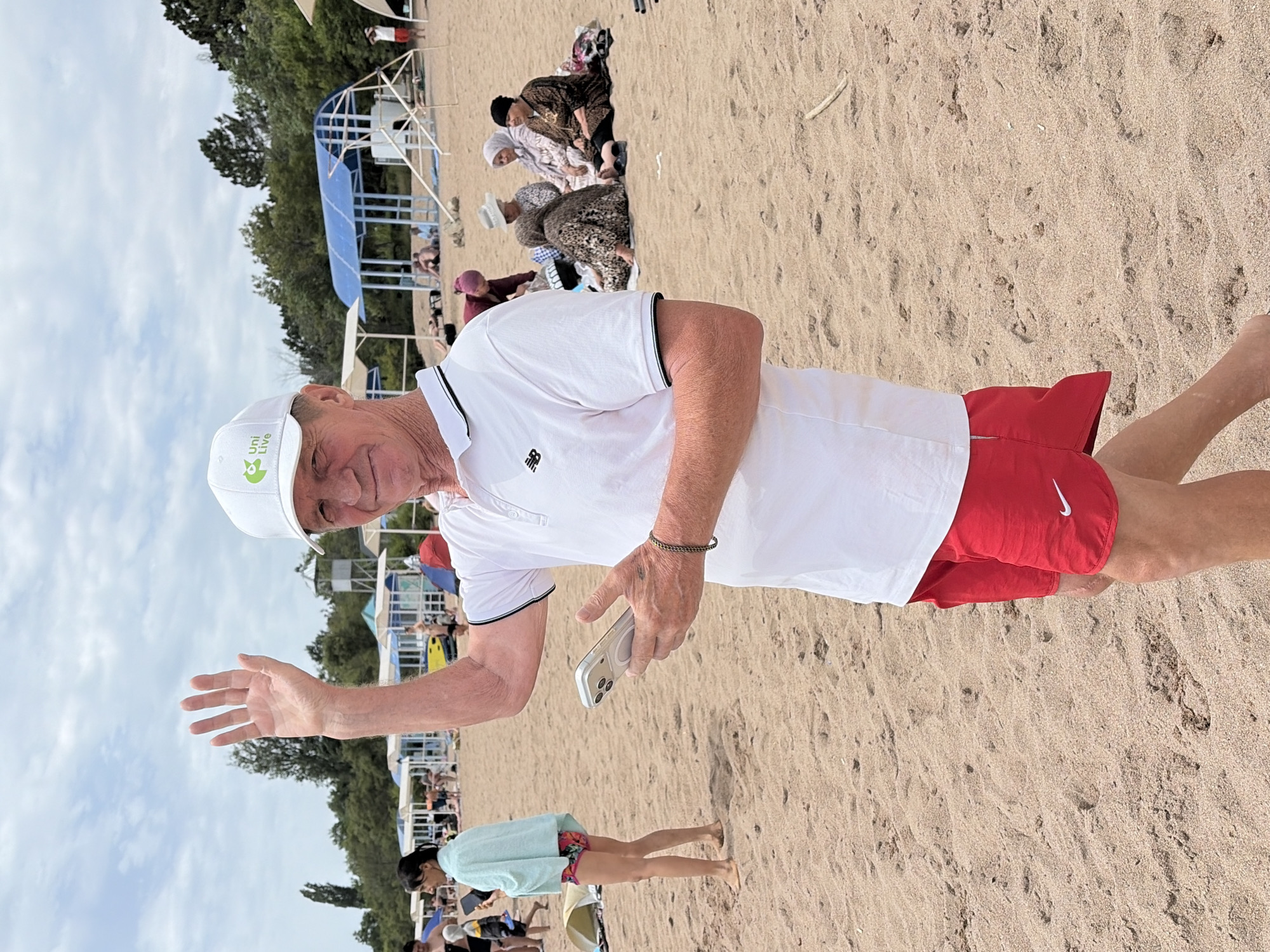 An elderly Caucasian male wearing a white polo shirt, red athletic shorts, and a white baseball cap with a logo stands on a sandy beach. He is facing the camera and waving with his left hand, which is raised. He holds a silver smartphone in his right hand. In the background, other beachgoers are visible, some sitting on blankets and others walking. There are beach umbrellas and some trees in the distance. The sky is overcast with some clouds.