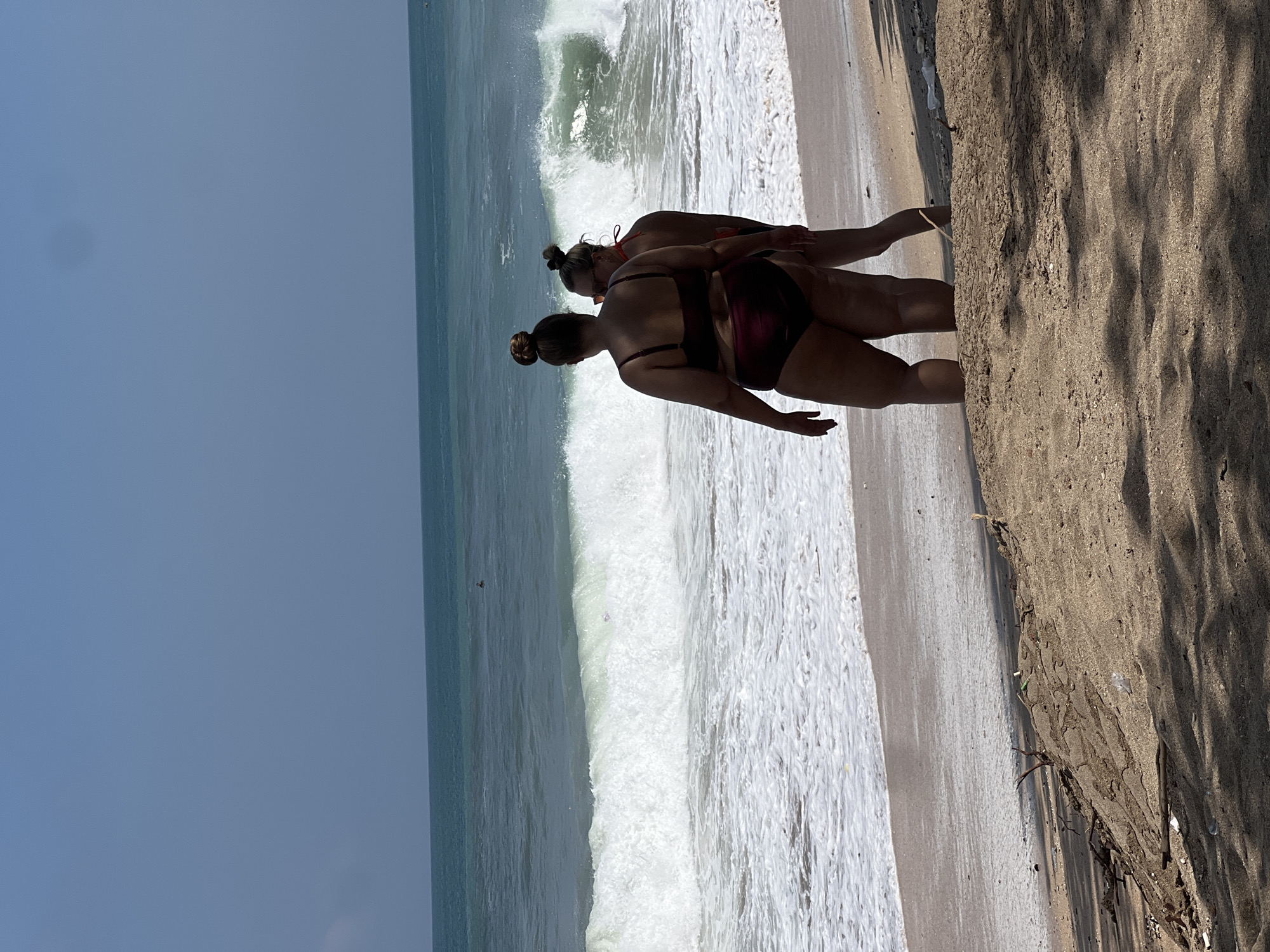 Two women are standing on a sandy beach in Kuta, Indonesia, facing the ocean. The woman in the foreground is larger and wears a dark red bikini, with her back to the camera. The second woman, smaller and on the right, is wearing a red bikini top and appears to be looking out at the ocean. Large waves are crashing on the shore. The sky is clear and blue, and the water is a deep turquoise. In the distance, a person can be seen in the water. The foreground of the image is dominated by sand and shadows, with some debris and vegetation visible.