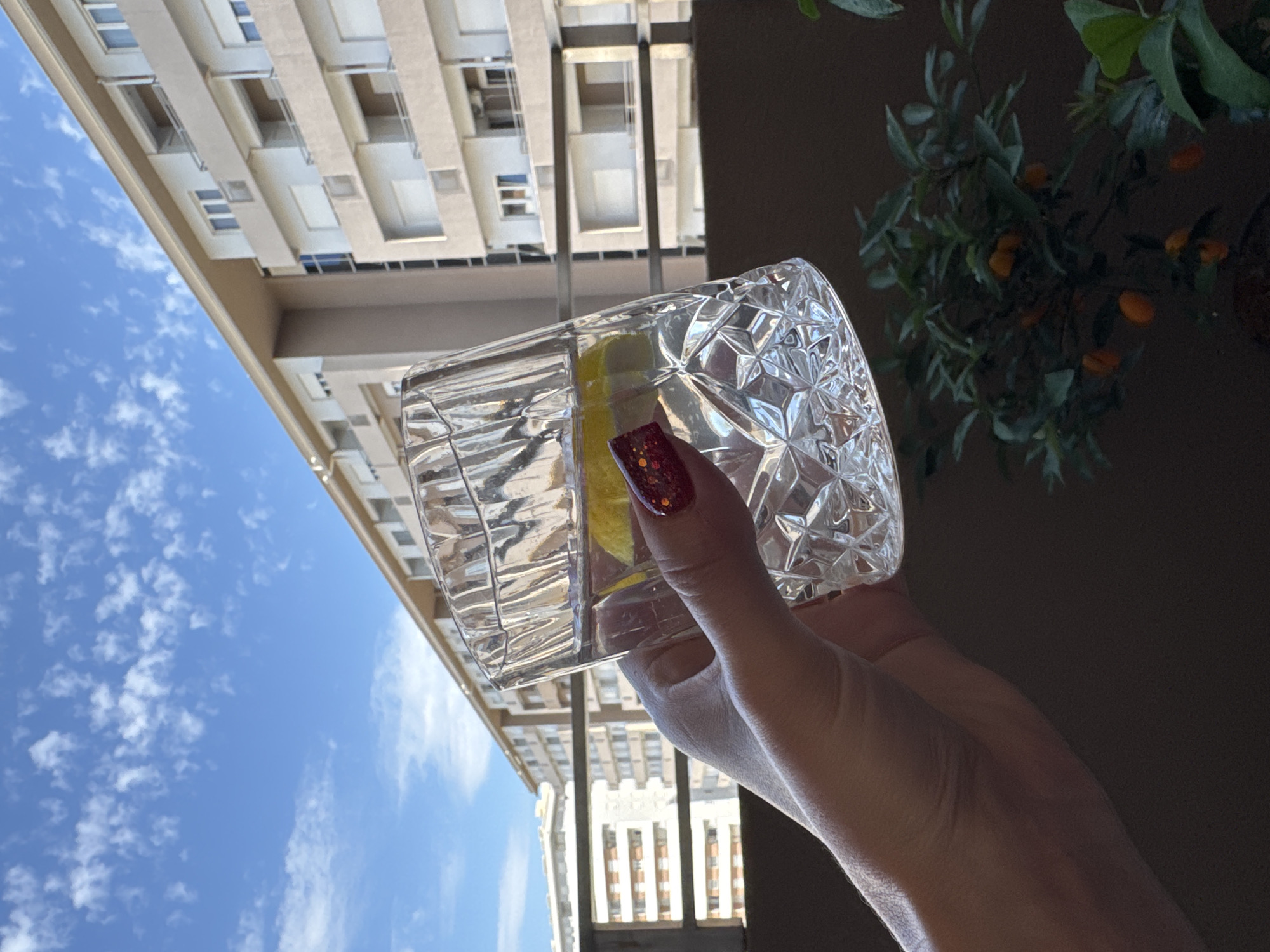 A person's hand, with glittery red nail polish, holds a crystal glass filled with a clear liquid and a slice of lemon. The glass is positioned in front of a backdrop of buildings under a bright blue sky with scattered clouds. In the foreground, a small tree with orange fruits is visible. The scene appears to be on a balcony or terrace.