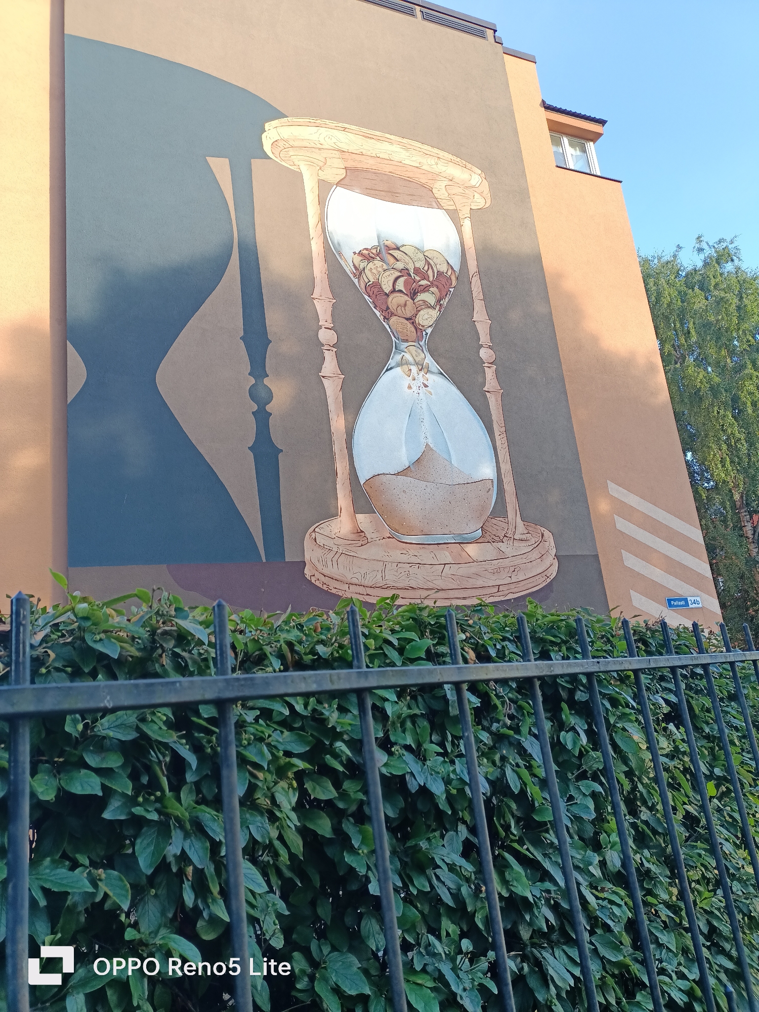 The image shows a mural painted on the side of a building in Tallinn, Estonia. The mural depicts an hourglass filled with coins. In the foreground, a black metal fence is visible in front of a dense green hedge. The building has a beige facade with some abstract shapes painted on it. The sky is blue and clear, and trees are visible on the right side of the image. A small sign on the building reads "Paljassaare 34b".