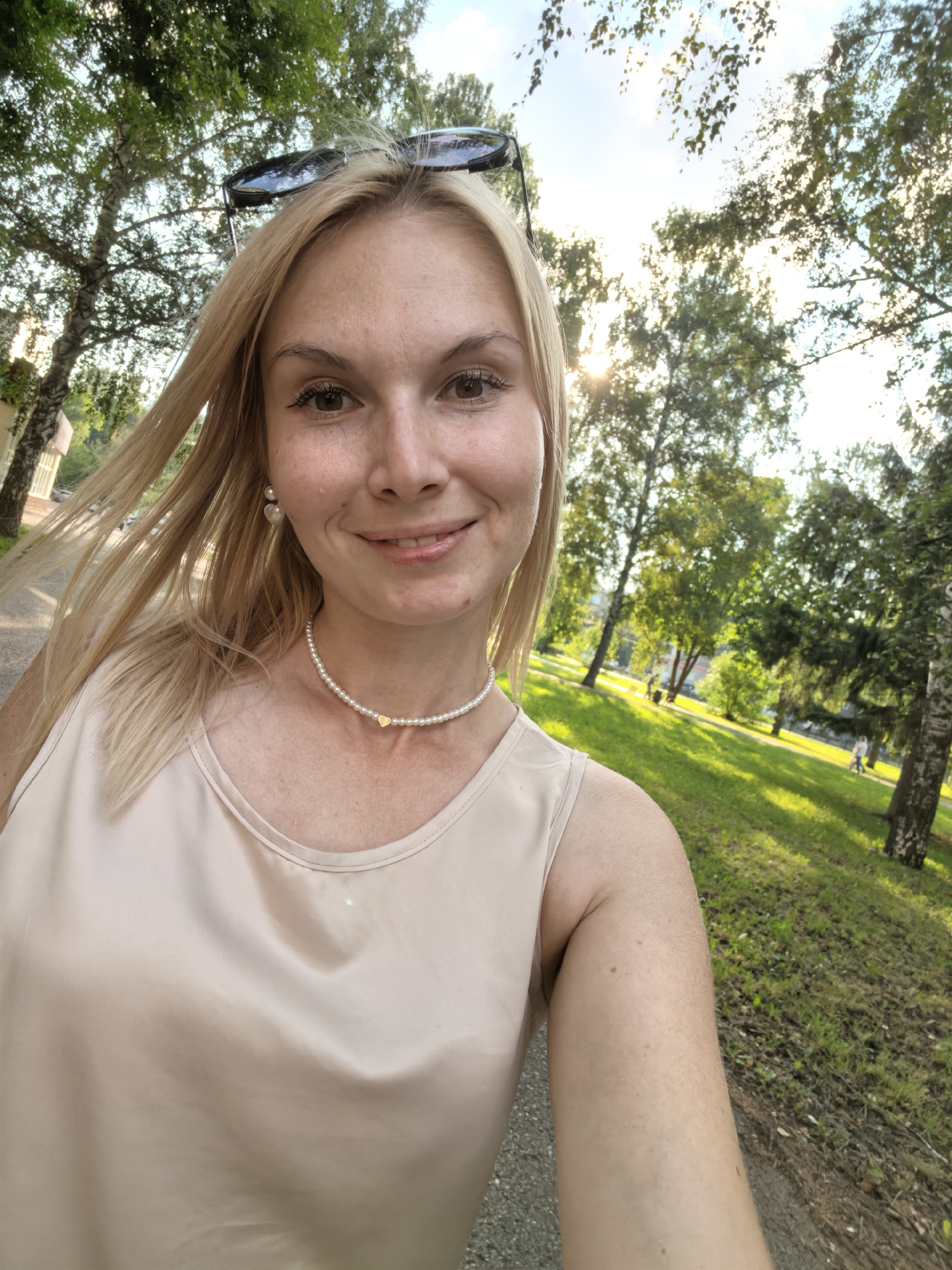 A blonde woman with sunglasses perched on her head is taking a selfie in a park. She is wearing a light beige tank top and a pearl necklace with a gold heart pendant. Her hair is blowing in the wind. The background shows trees, a grassy area, and a path. The sun is shining through the trees.