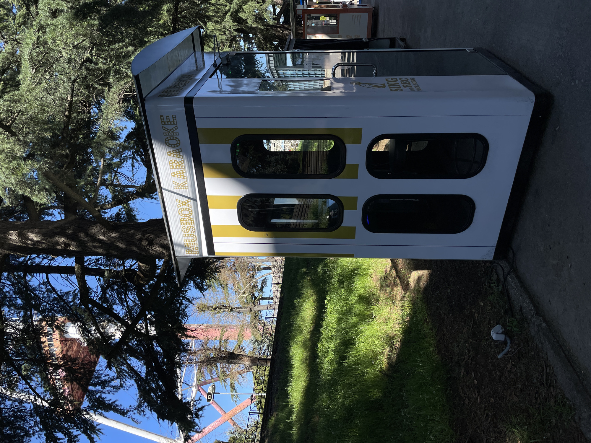 The image shows a "Husky Karaoke Box" in Tbilisi, Georgia. It is a white and yellow karaoke booth with four rectangular windows. The booth stands on a concrete surface, with a grassy area and trees behind it. In the background, there is a Ferris wheel visible through the trees.