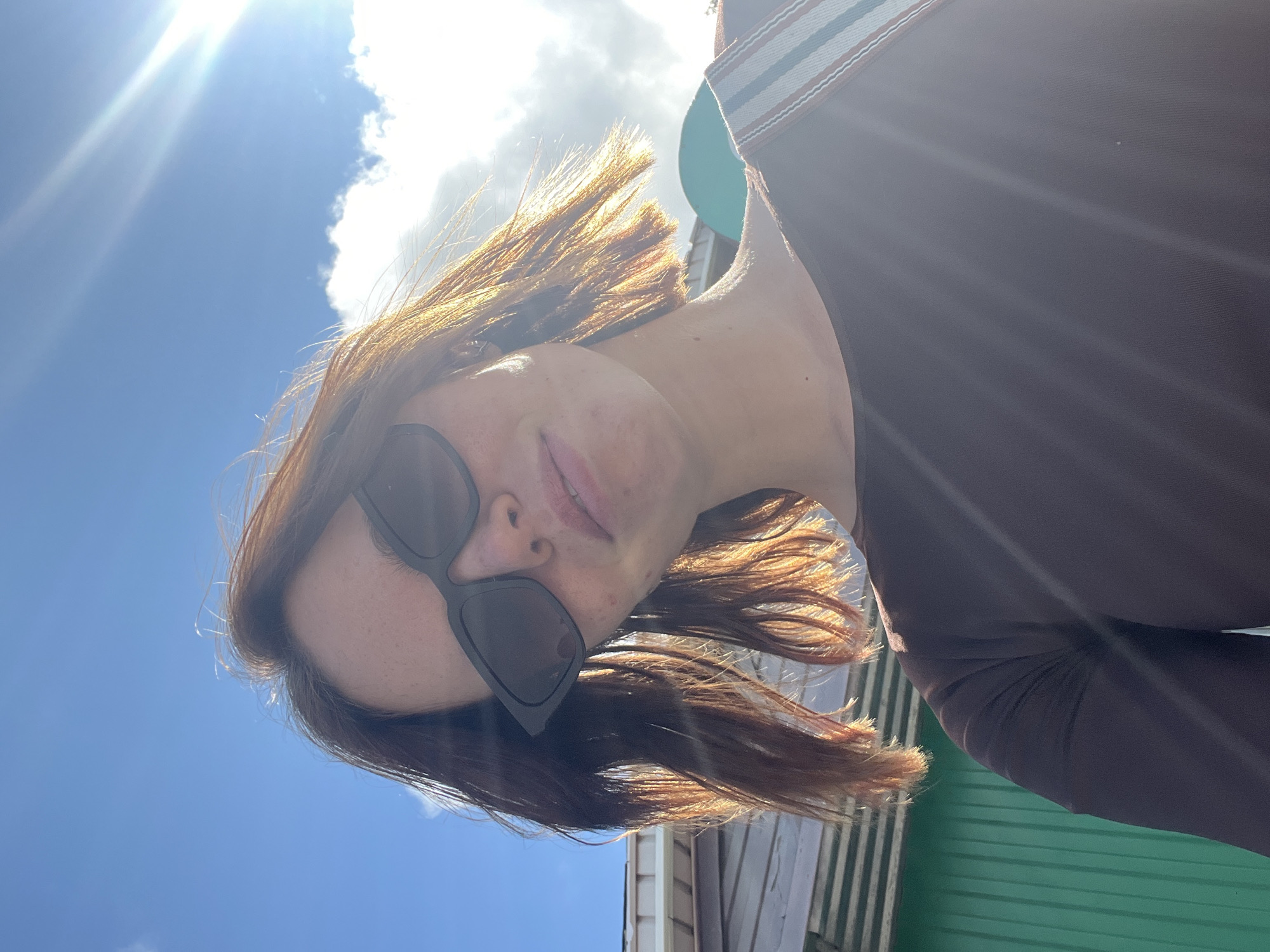 A young woman with shoulder-length brown hair, wearing dark sunglasses and a brown long-sleeved shirt, poses for a selfie outdoors. She is standing in front of a building with light-colored siding and a green corrugated metal wall. The sky above is bright blue with scattered white clouds, and the sun is shining brightly, creating lens flare and illuminating her hair with a warm glow. Her expression is relaxed, and she is looking towards the camera. A portion of a striped strap, possibly from a bag, is visible over her shoulder. The image appears to be taken on a sunny day. There is no indication of the specific location within Chaikovsky, Russia.