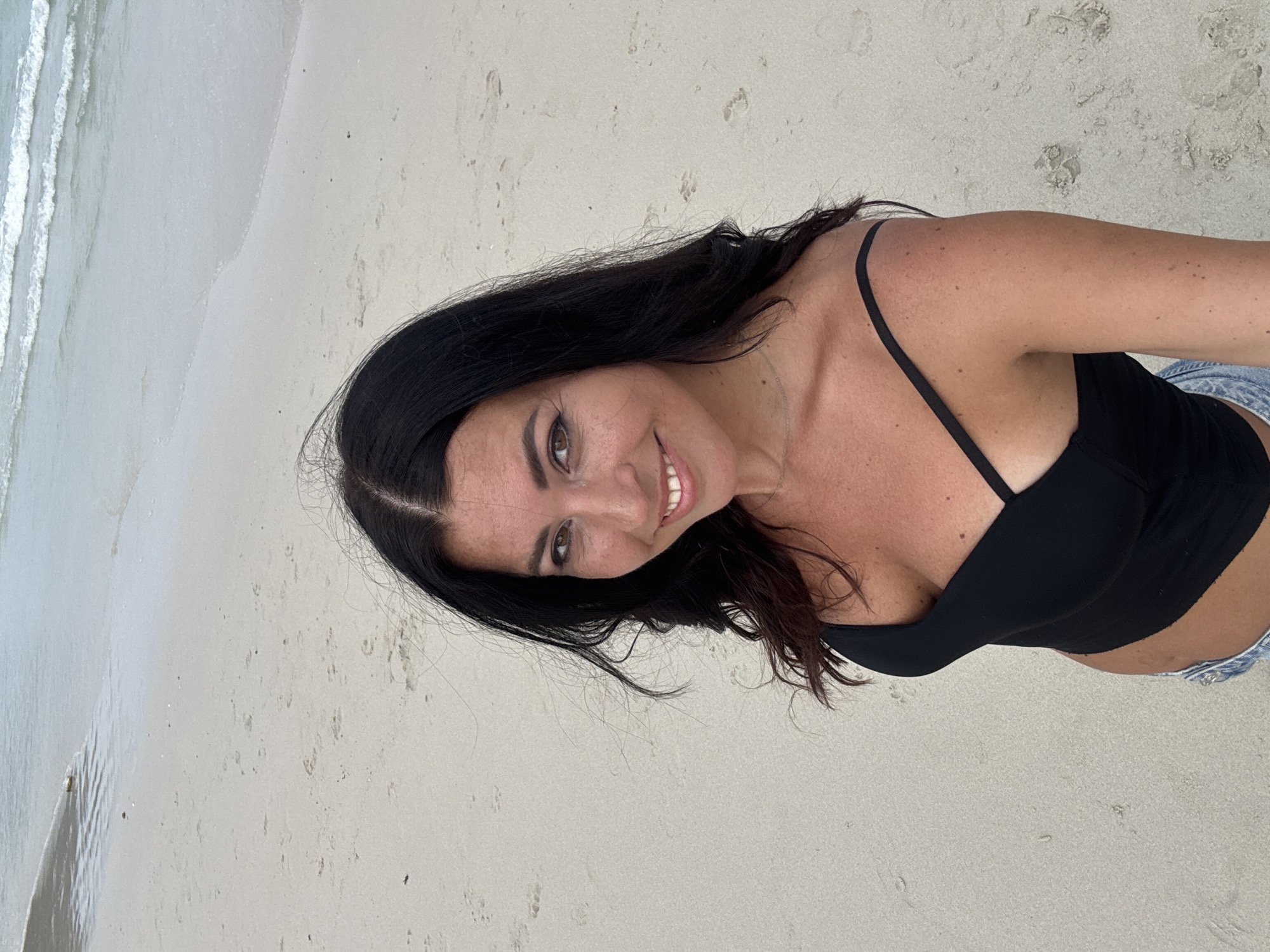 A woman with dark hair is standing on a sandy beach with waves visible in the background. She is wearing a black top and denim shorts. The scene is likely on a beach, possibly in Ban Haad Rin, Thailand.