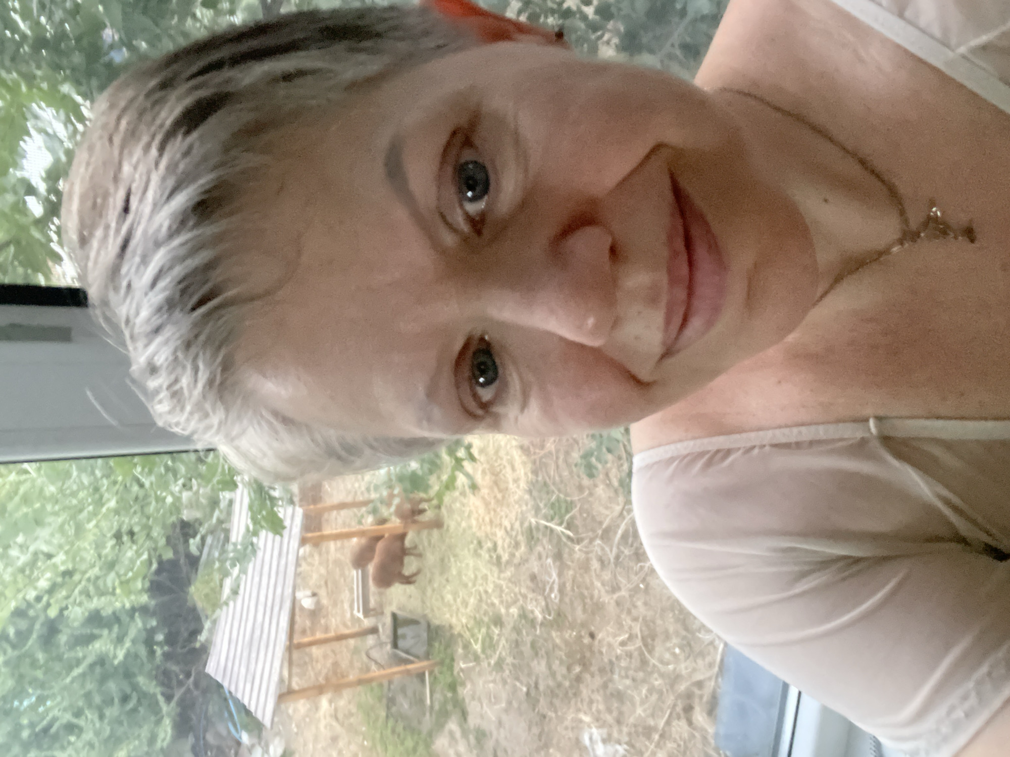 A selfie of a woman with short gray hair is taken indoors, with a blurred background of greenery and a small wooden structure. Two brown animals, possibly sheep or goats, are visible behind the woman, near the structure. The woman is wearing a light-colored, sheer top and a necklace. The setting appears to be a backyard or a rural area, but no specific location in Almaty, Kazakhstan can be identified from the image.