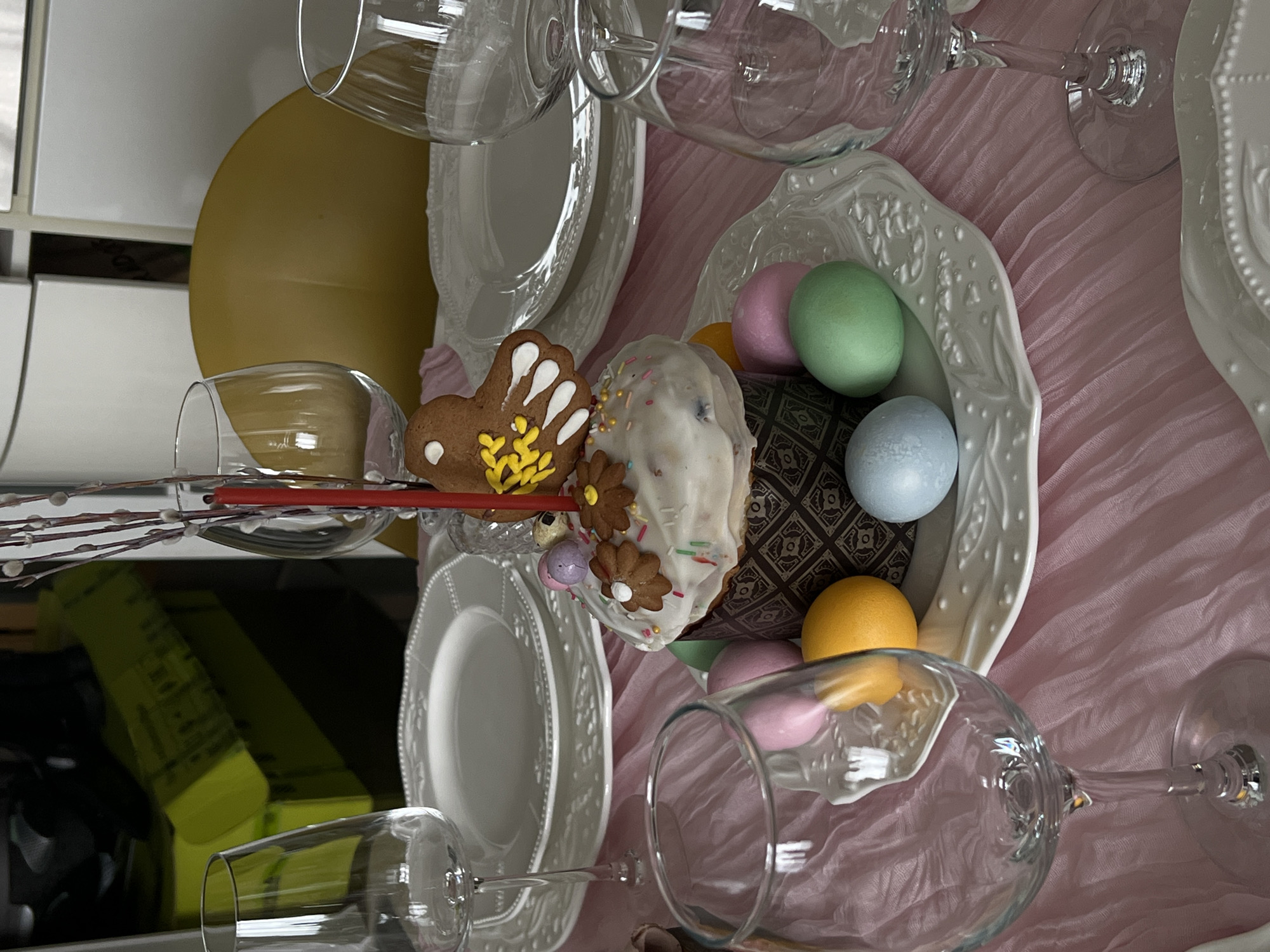 A tabletop setting for a celebration, possibly Easter, is oriented on its side. There's a light pink, textured tablecloth. Multiple clear, stemmed wine glasses are scattered around, some lying on their sides. Several white, decorative plates are stacked. A shallow, white, oval bowl contains colorful Easter eggs in shades of green, blue, pink, and yellow. Next to the bowl is a decorative gingerbread cookie shaped like a butterfly and a small cake covered in white frosting with sprinkles and decorative chocolate flowers. The items are close together, giving a sense of abundance.
There is no indication of the location in the city of Khimki, Russia.