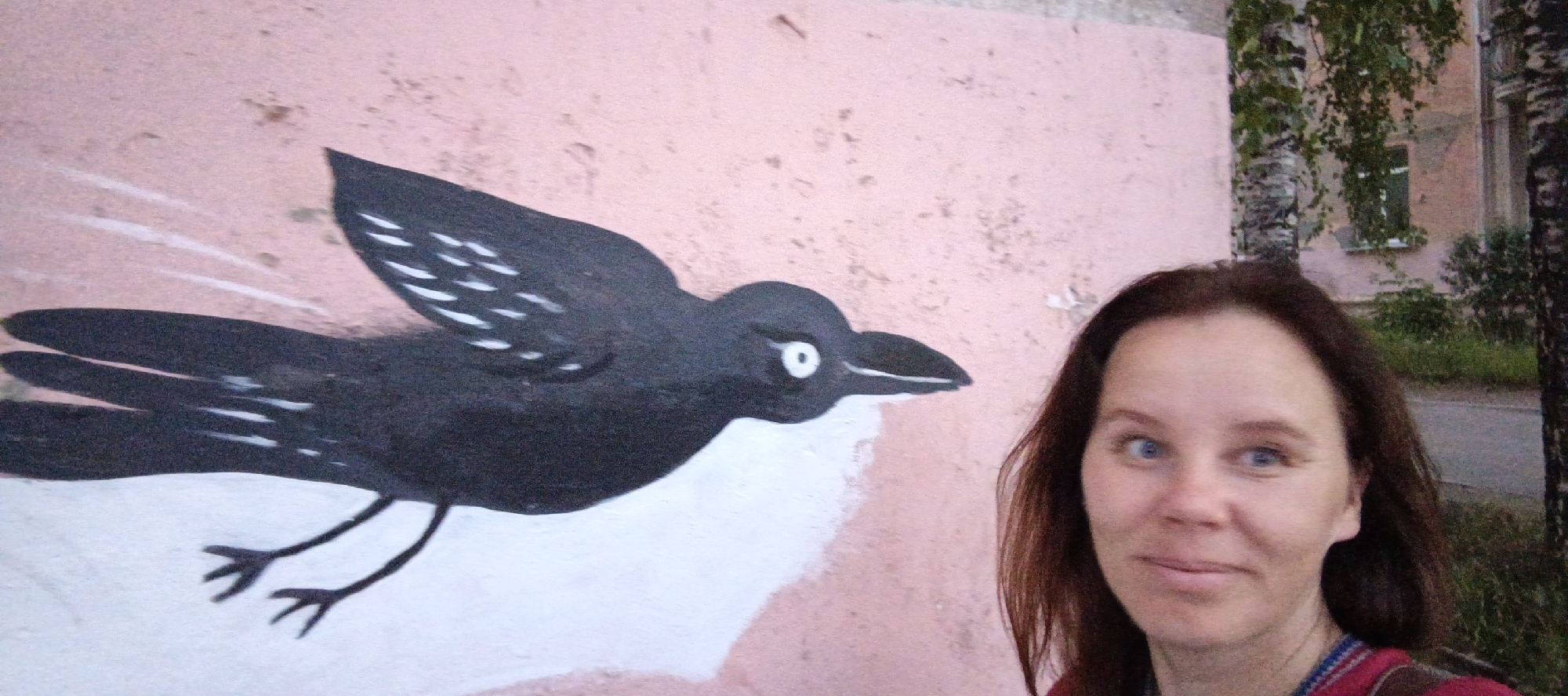 In Votkinsk, Russia, a woman with brown hair and blue eyes stands in front of a mural of a black bird on a pink wall. The bird appears to be in flight, with its wings spread and white markings on its feathers. The woman is smiling and looking at the camera. Behind the mural, a tree with white bark and green leaves is visible, along with a building. The overall scene appears to be an outdoor public space.
