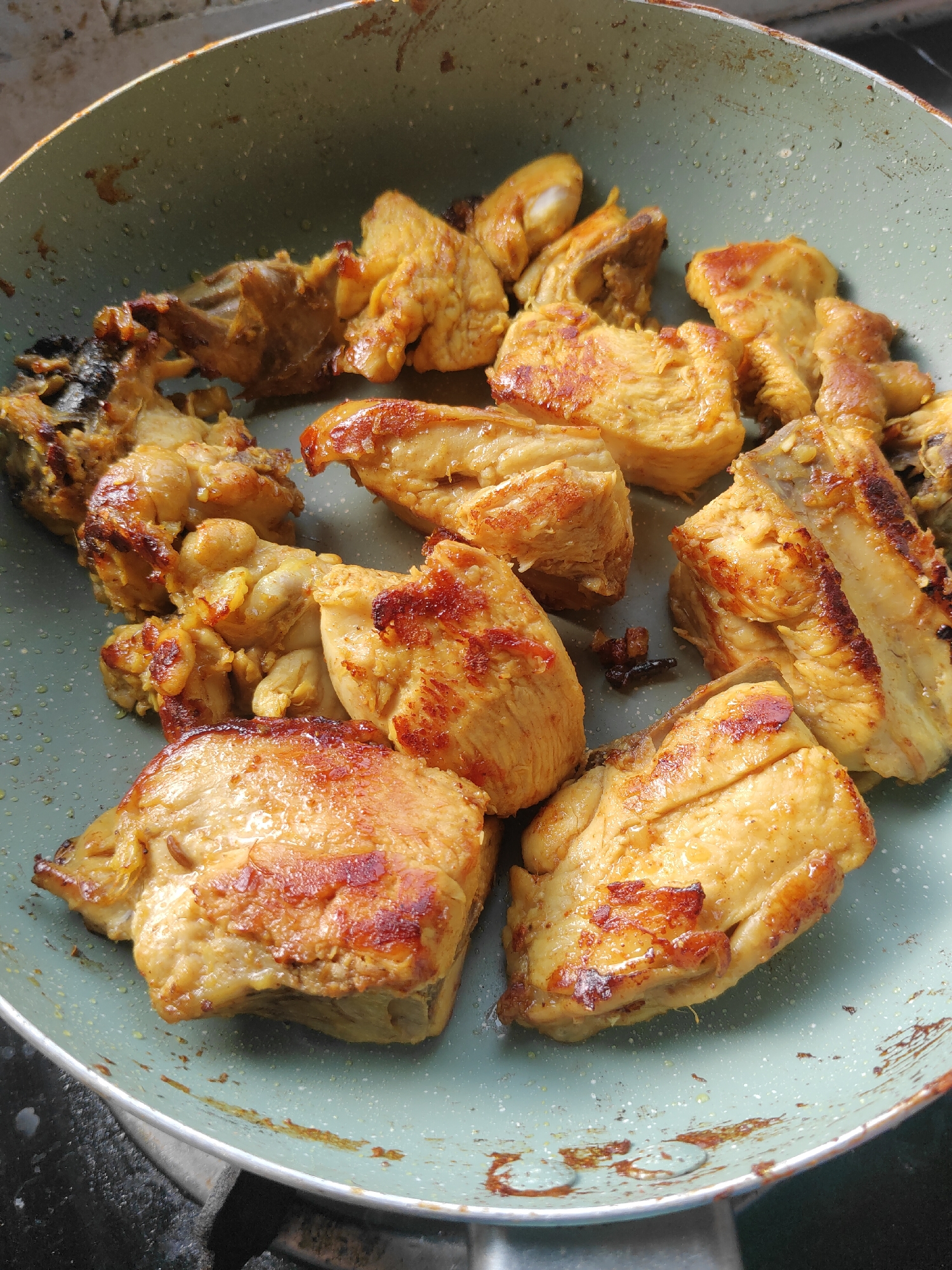 The image shows chicken pieces being cooked in a pan. The chicken is golden brown and appears to be seasoned. Some pieces are slightly charred, indicating they are well-cooked. The pan is light green with a speckled interior and is placed on a dark surface, possibly a stove. There is some residue or discoloration on the pan's surface and rim. The scene is an indoor kitchen setting, consistent with a home in Watesnegoro, Indonesia, where such a meal might be prepared. There are no people visible in the image.