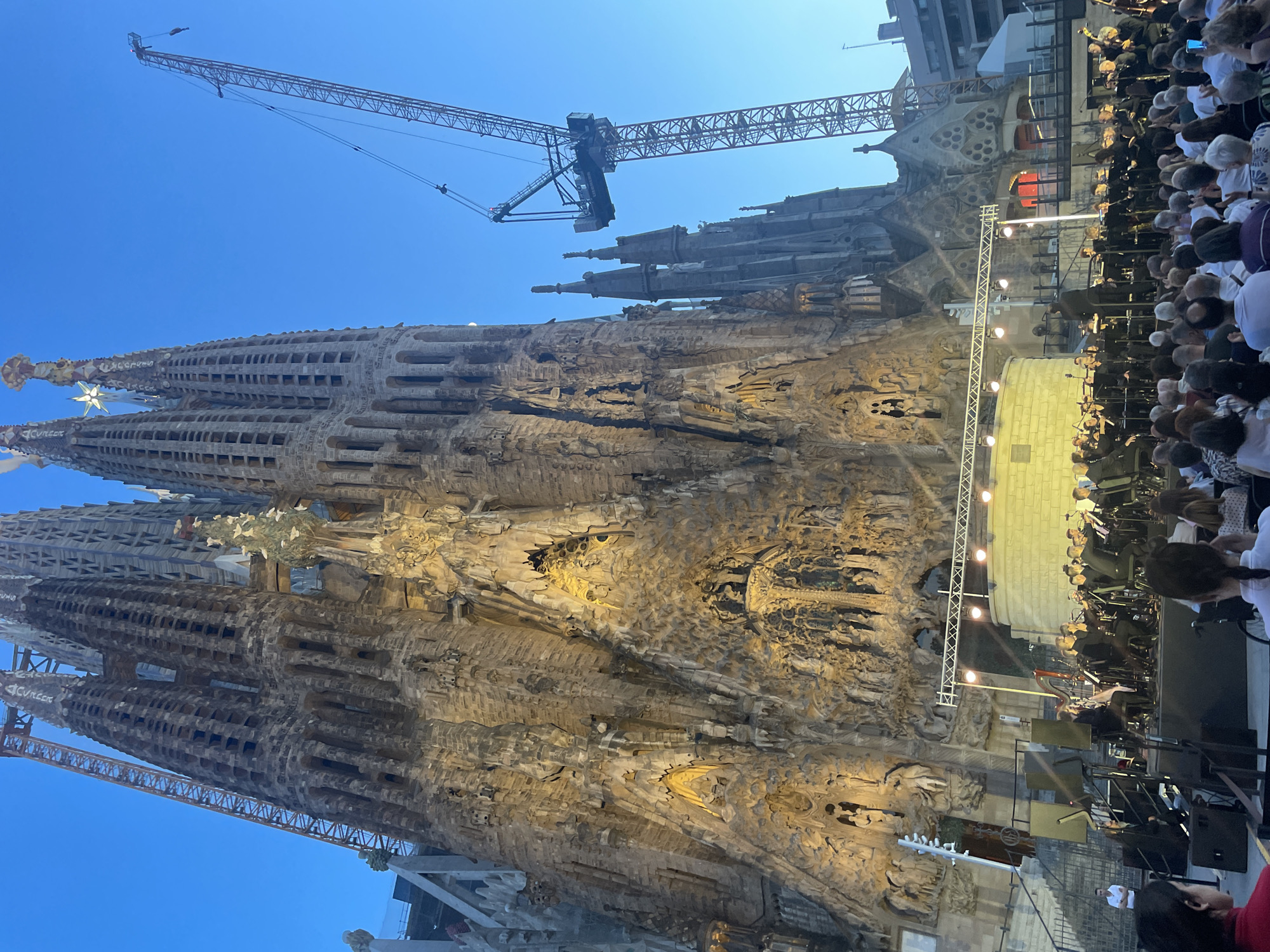 This image depicts an outdoor orchestral performance in front of the Sagrada Familia basilica in Barcelona, Spain. The basilica, a UNESCO World Heritage Site, dominates the background with its intricate facade illuminated by warm lights. Two large construction cranes are visible, one on the left and another on the right, indicating ongoing construction or restoration work on the iconic structure. In the foreground, a symphony orchestra is set up on a stage with a lighting rig, performing for a seated audience. The sky is a clear, twilight blue. The scene captures a cultural event taking place at a significant architectural landmark.
