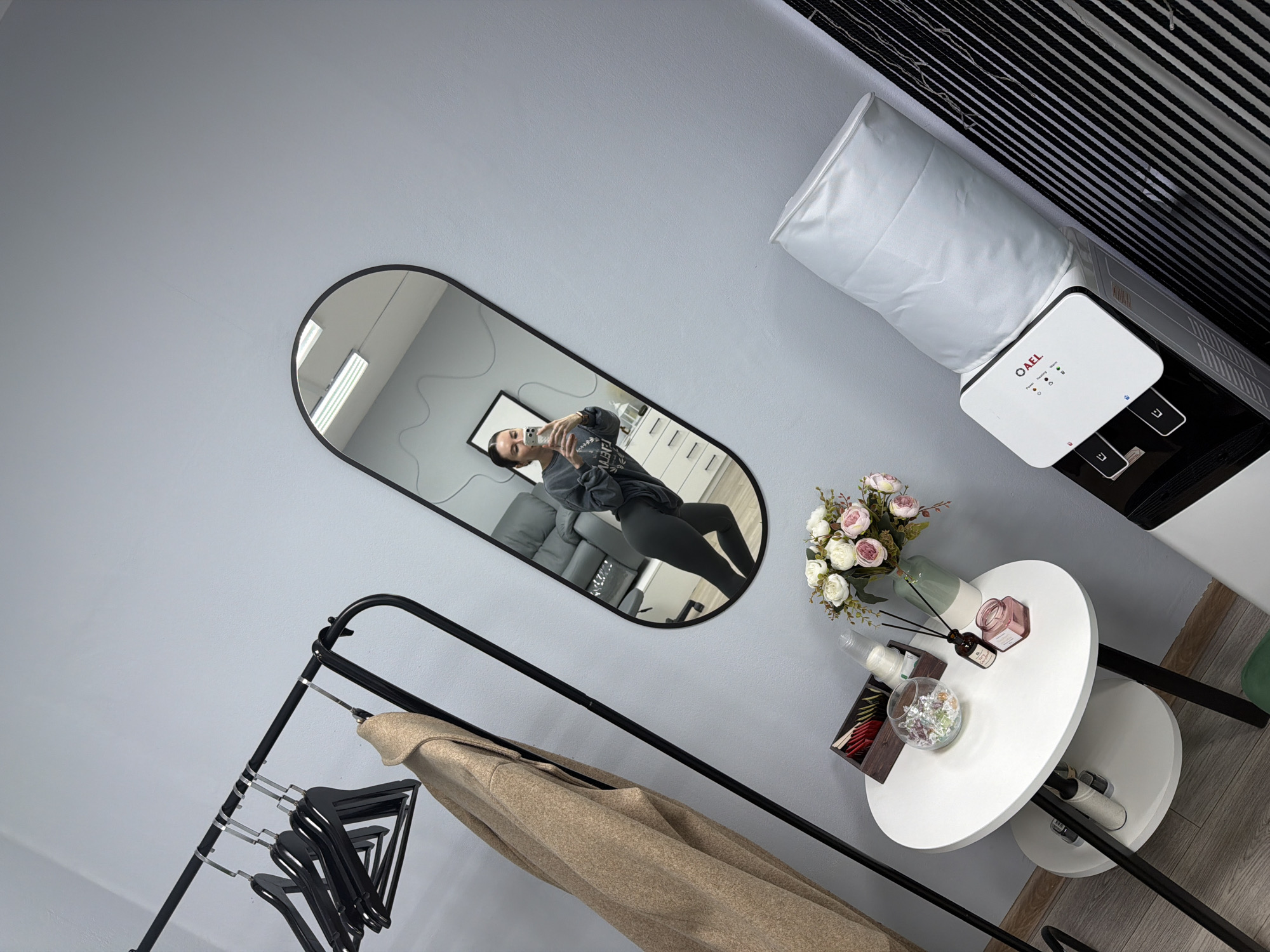 The image captures an indoor scene, likely in Perm, Russia, featuring a modern and functional aesthetic. The primary subject is a woman visible in the reflection of a large, black-framed oval mirror, taking a selfie with her phone.
The room has light grey walls. In the foreground, a black metal clothing rack holds several black empty hangers and a beige or light brown coat. To the right of the mirror, a white two-tiered round table is decorated with a vase of artificial pink and white flowers, a reed diffuser, a pink jar candle, a glass bowl containing small candies or decorative stones, and a wooden box holding tea bags.
Further to the right and slightly behind the table, a white and black "O AEL" brand water cooler/dispenser stands against the wall, topped with a white fabric cover over the water bottle. Black horizontal blinds or slats are visible in the background, above the water cooler. The floor appears to be a light wood or laminate.
The activity taking place is the woman photographing herself in the mirror. The lighting suggests daytime, likely from artificial sources like the fluorescent lights reflected in the mirror. Visible text includes "O AEL" on the water cooler, along with small red, green, and blue indicator lights. Mirrored text, possibly Cyrillic, is visible on the woman's dark sweatshirt, appearing as "ЧETVІ".